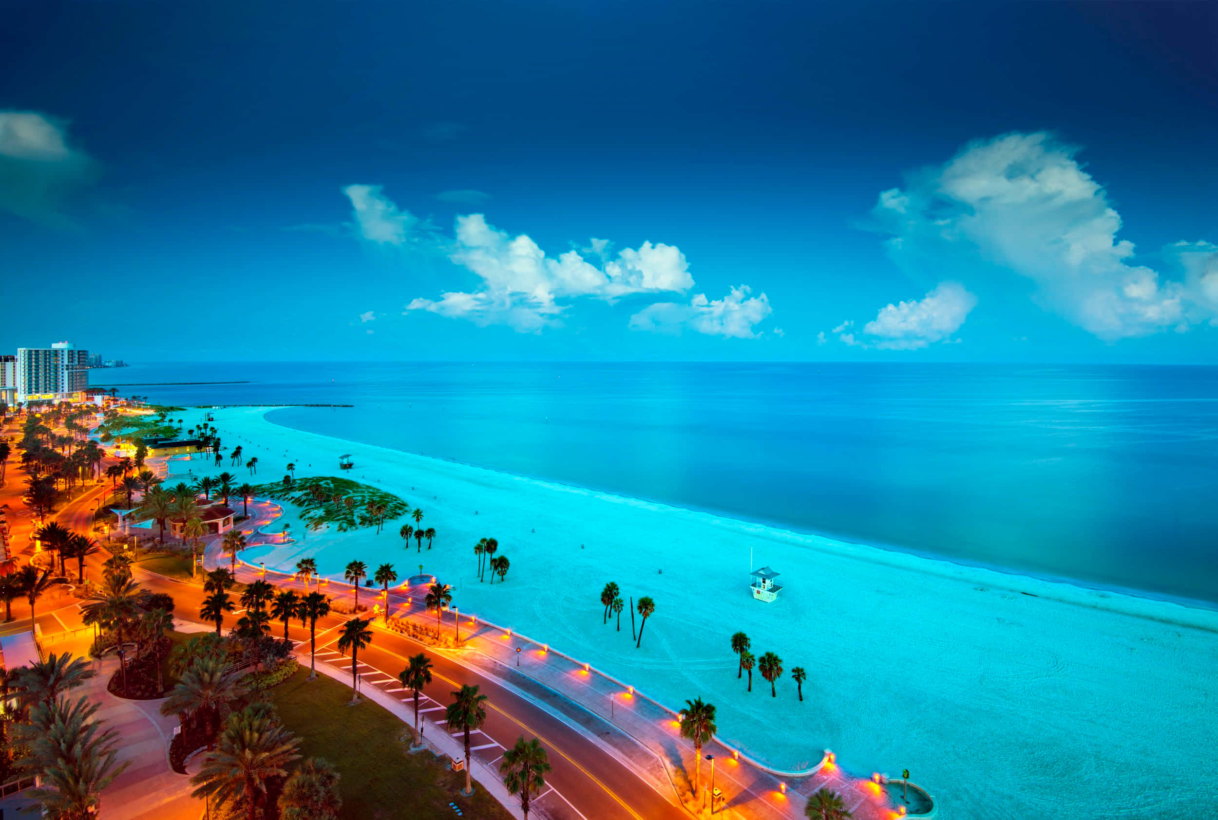 Beautiful Beach 桌布 壁紙 — 充滿活力的黃昏海灘場景，在深藍色天空和蓬鬆雲朵下，橙色街燈沿著棕櫚樹排列在碧綠海水旁閃閃發光。