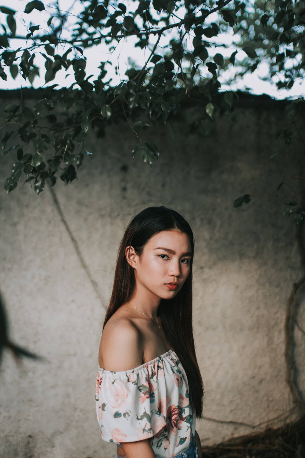 年輕女性穿花卉上衣站在混凝土牆附近的樹枝下
