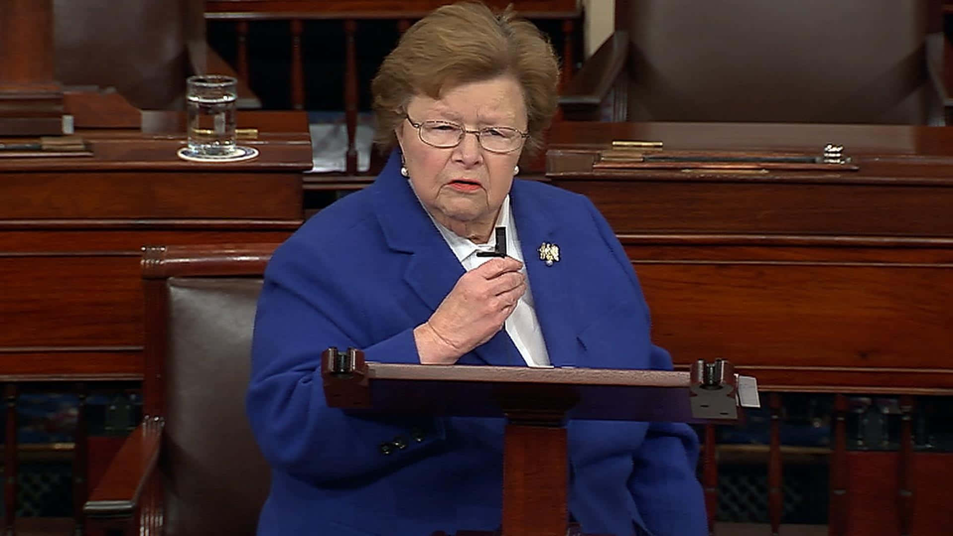 先驅女性政治家 Barbara Mikulski 壁紙 — 一位身穿藍色套裝的老年女性在正式立法會議廳的木製講台上發言。