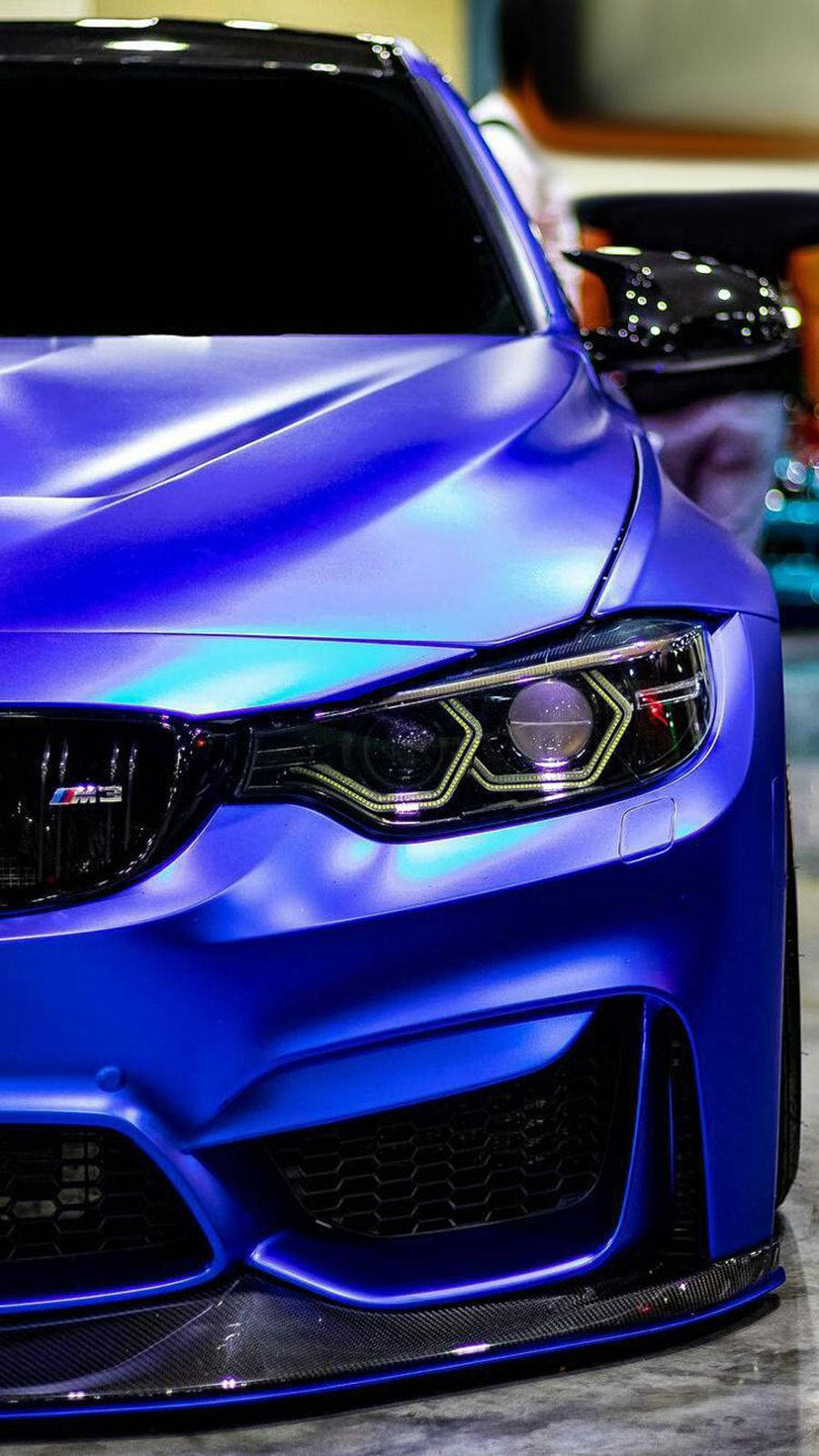 光澤亮麗的藍色BMW 壁紙 — 啞光藍色BMW M3嘅特寫，配備發光頭燈同碳纖維飾件，喺明亮嘅室內燈光下。