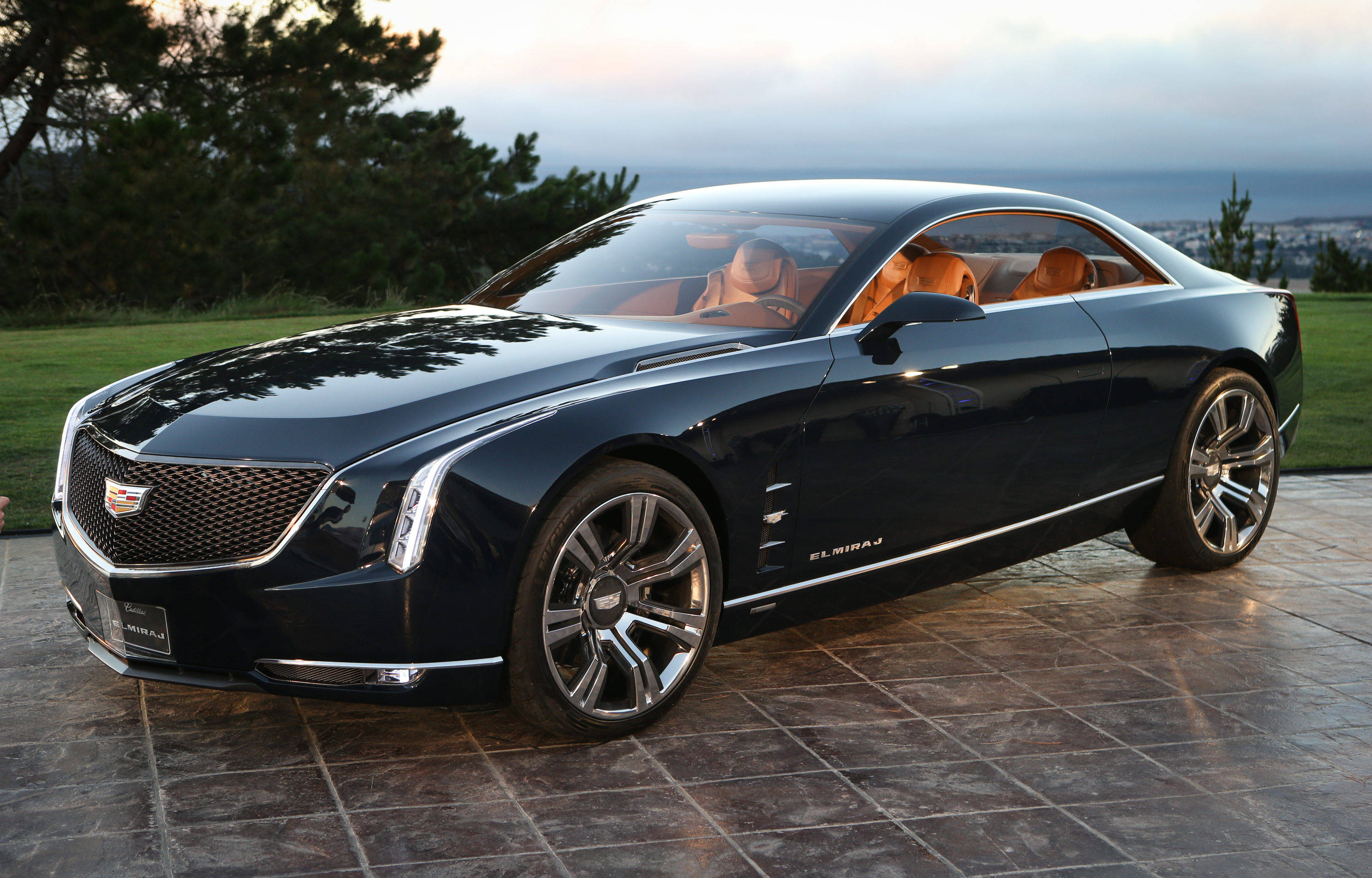光澤黑凱迪拉克 壁紙 — 一輛時尚的深藍色 Cadillac Elmiraj 豪華轎跑車停在石磚上，背景是草地山坡和遠處的城市景觀，正值黃昏。