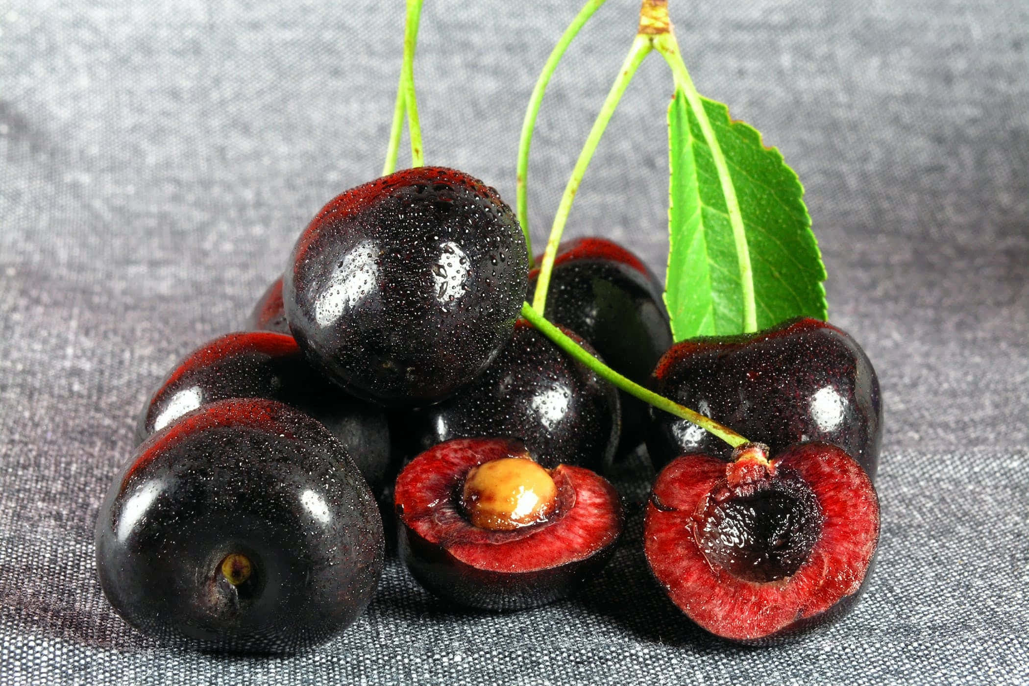 Black Cherry 桌布 壁紙 — 光澤黑色車厘子帶有鮮紅色內部及綠色莖部，放在有紋理的灰色布料上。