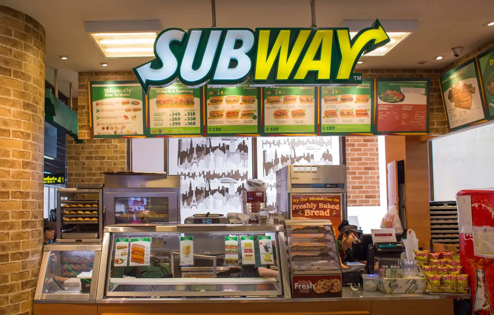 Subway 桌布 壁紙 — 光線充足的Subway三文治店內部，設有磚牆、菜單板及裝滿新鮮烘焙食品的玻璃陳列櫃。