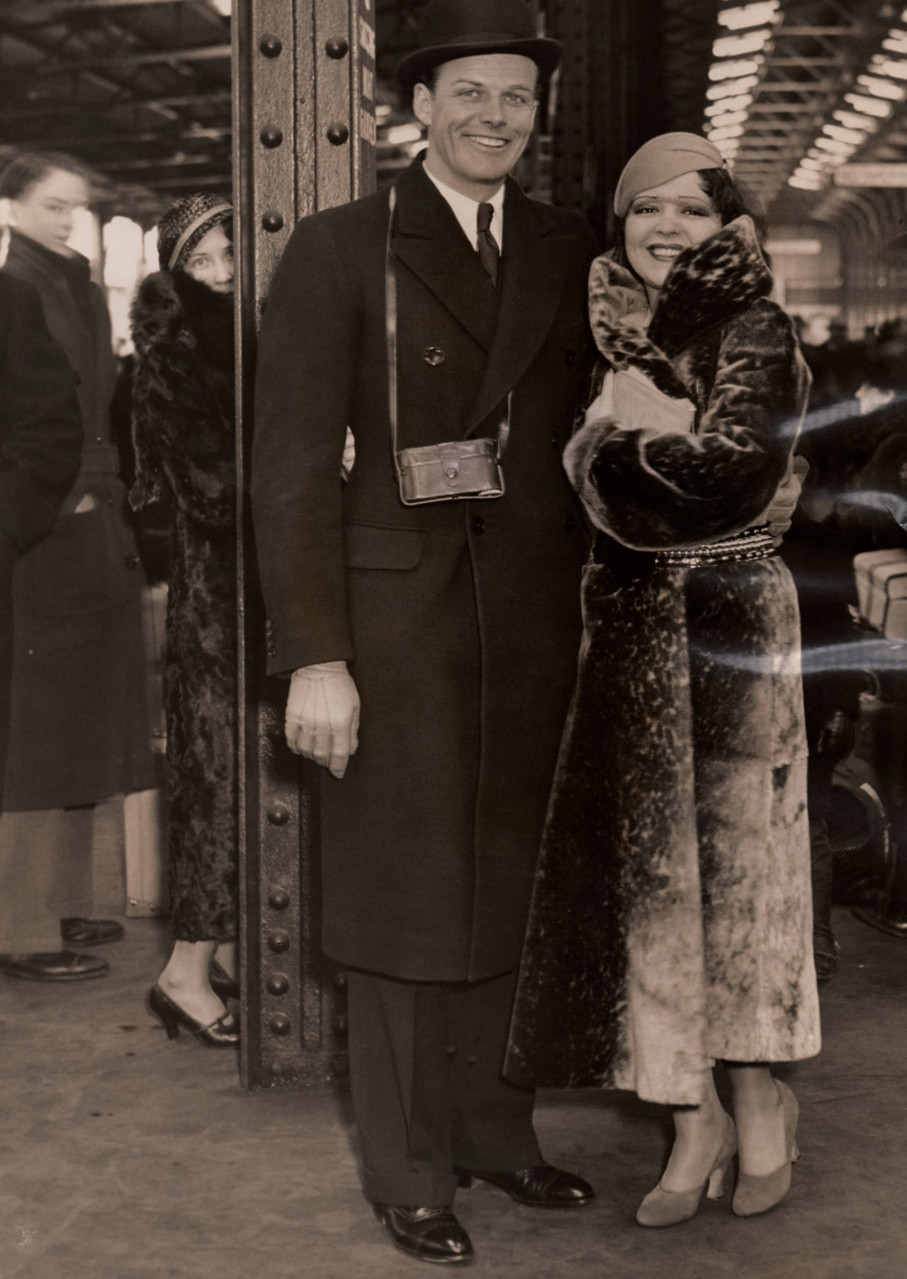 克拉拉·鮑與政壇丈夫 壁紙 — 一對微笑的1920年代夫婦穿著毛皮大衣和帽子，在繁忙的火車站拍照留念。