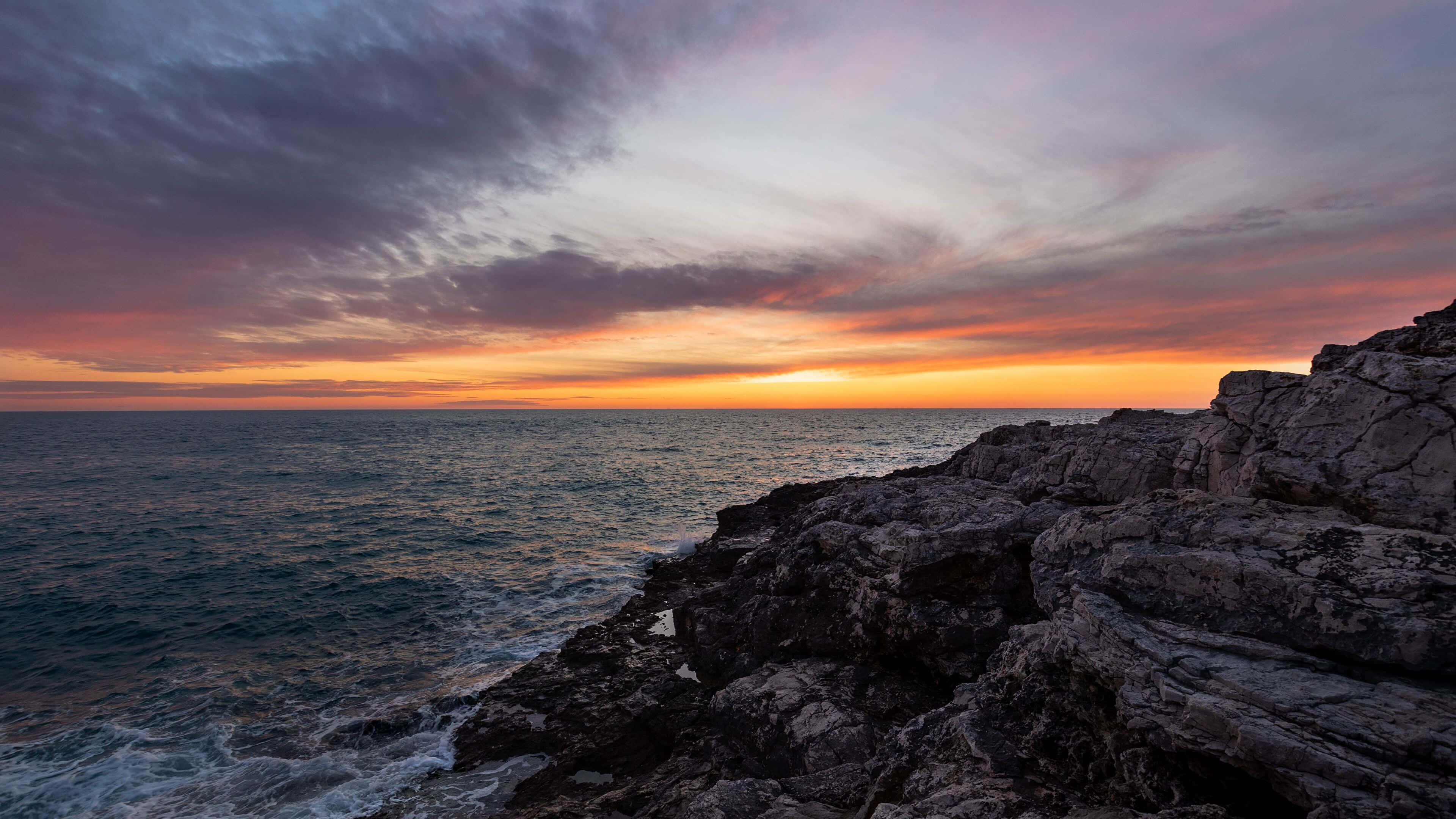 克羅地亞海岸的壯麗海浪 壁紙 — 戲劇性的夕陽將天空染成火熱的橙色和紫色，海浪拍打著崎嶇的海岸岩石。