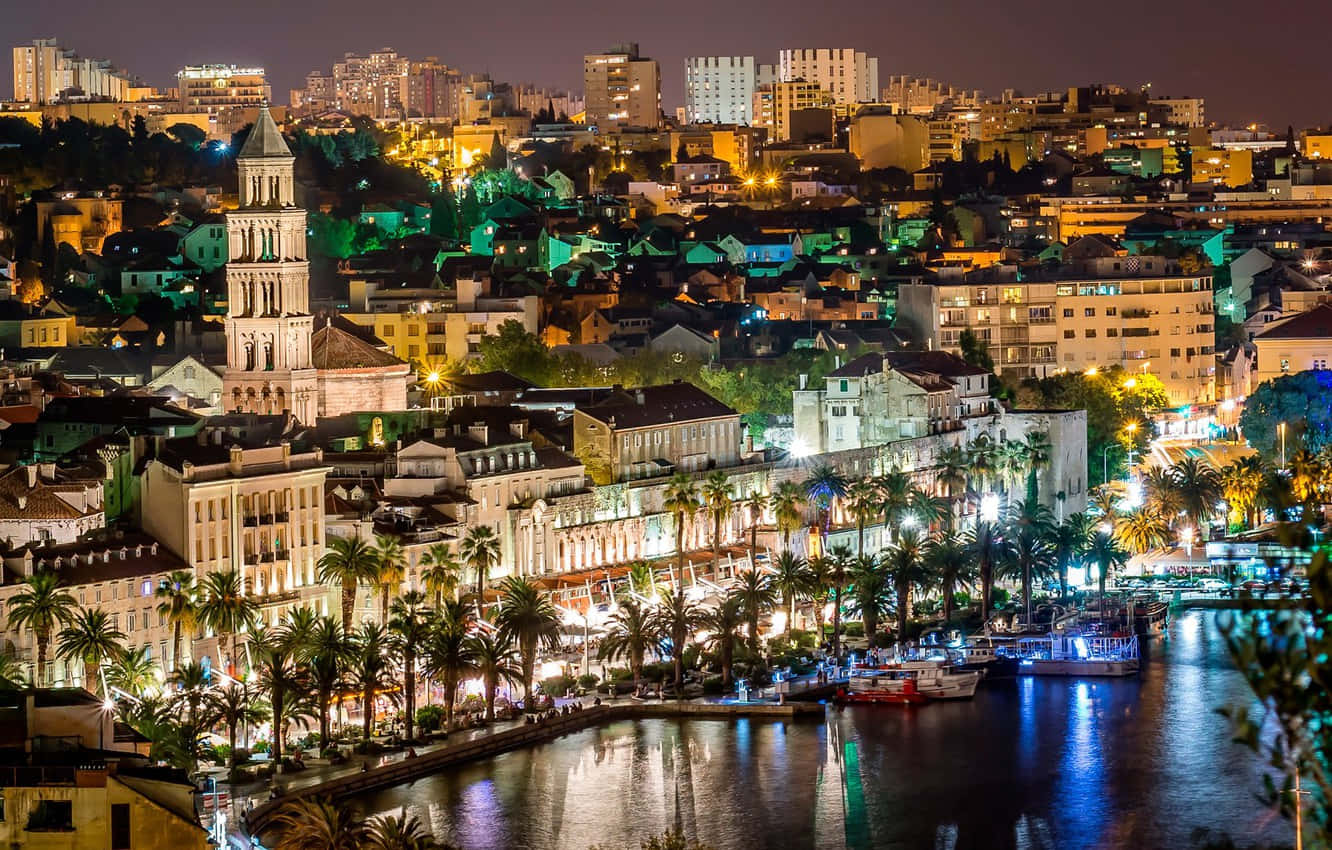 克羅地亞璀璨城市夜景 壁紙 — 充滿活力的夜景城市景觀，發光的建築物和棕櫚樹排列在反射性的海濱運河旁。