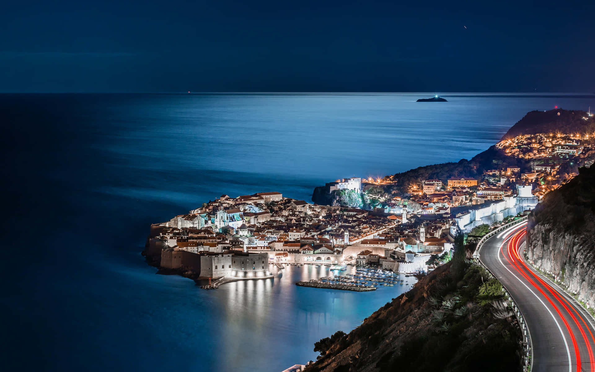 克羅埃西亞海濱夜景 壁紙 — 一個沿海小鎮在夜晚閃耀著溫暖的燈光，映照在平靜的藍色水面上，以及一條蜿蜒的道路上有紅色的汽車軌跡。