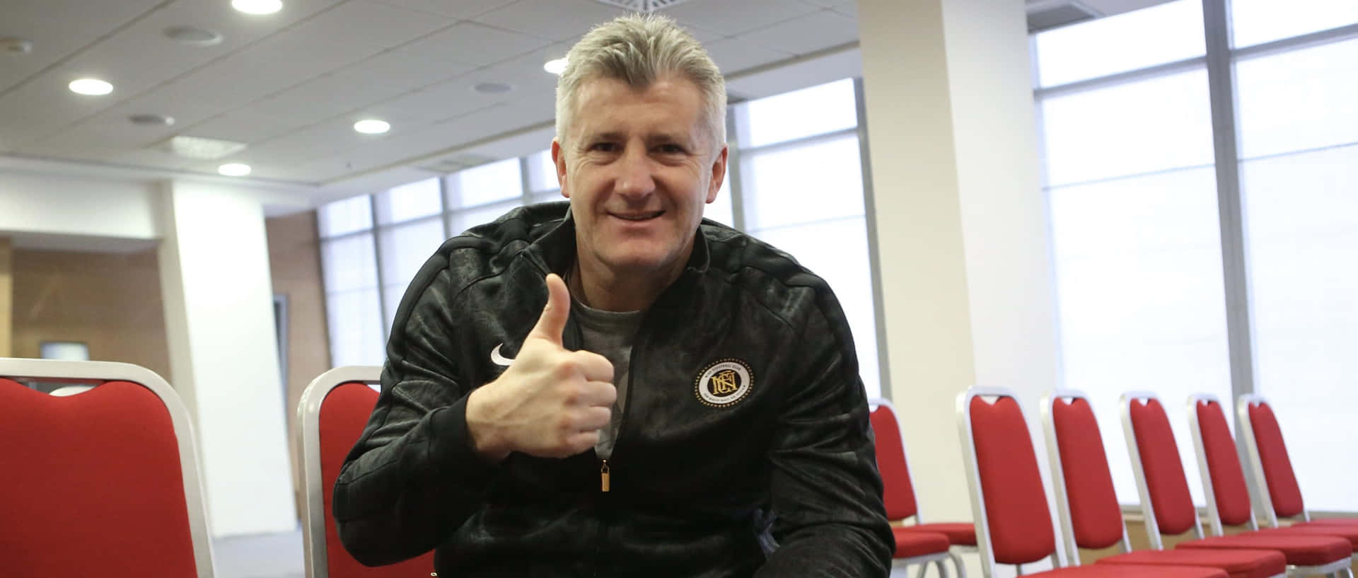 著黑色外套嘅男人喺有紅色椅嘅房入面豎起拇指