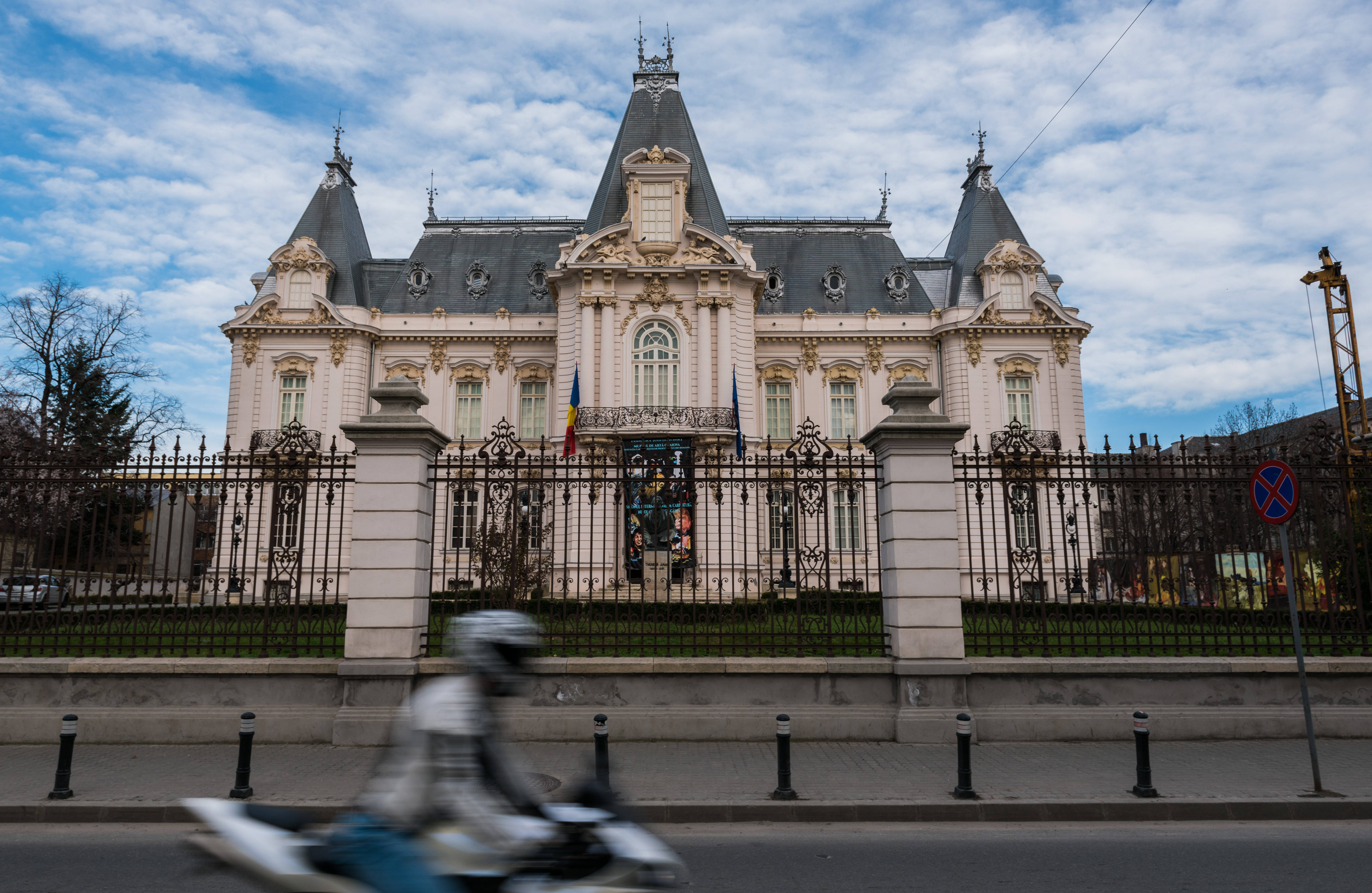 克羅瓦藝術博物館 壁紙 — 一座宏偉的裝飾性建築，灰色屋頂和白色外牆，矗立在鐵柵欄後面，一名電單車手在部分多雲的天空下於城市街道上疾馳而過。