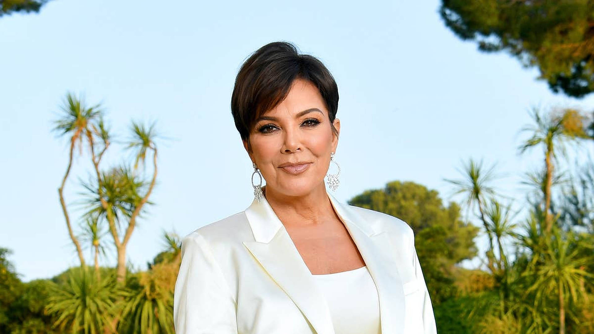 Kris Jenner 身穿白色西裝外套，在戶外微笑，身後有棕櫚樹。