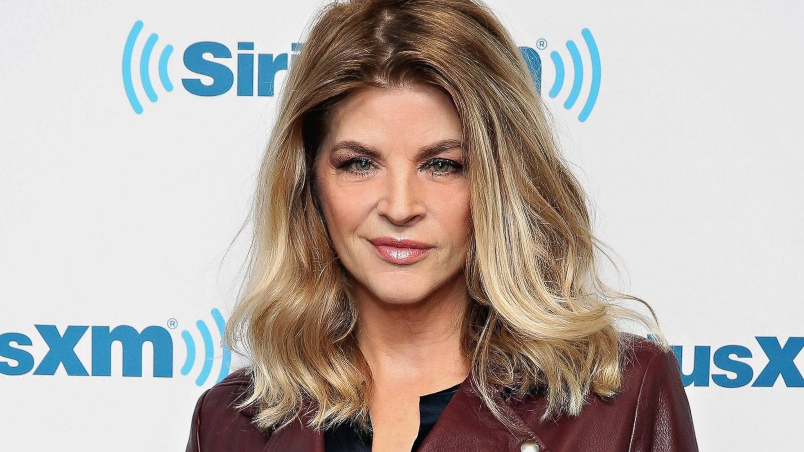 在SiriusXM活動中，身穿栗色外套的女性對著鏡頭微笑。