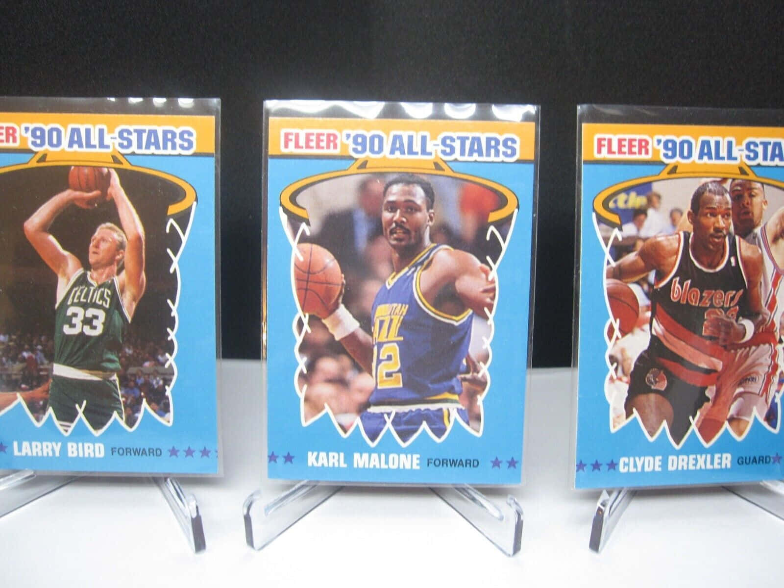 克萊德·德雷克斯勒、卡爾·馬龍與拉里·伯德 NBA 90年代全明星照片卡 壁紙 — 三張1990年Fleer All-Stars籃球球星卡， featuring Larry Bird、Karl Malone和Clyde Drexler，直立展示在透明支架上，背景為深色。