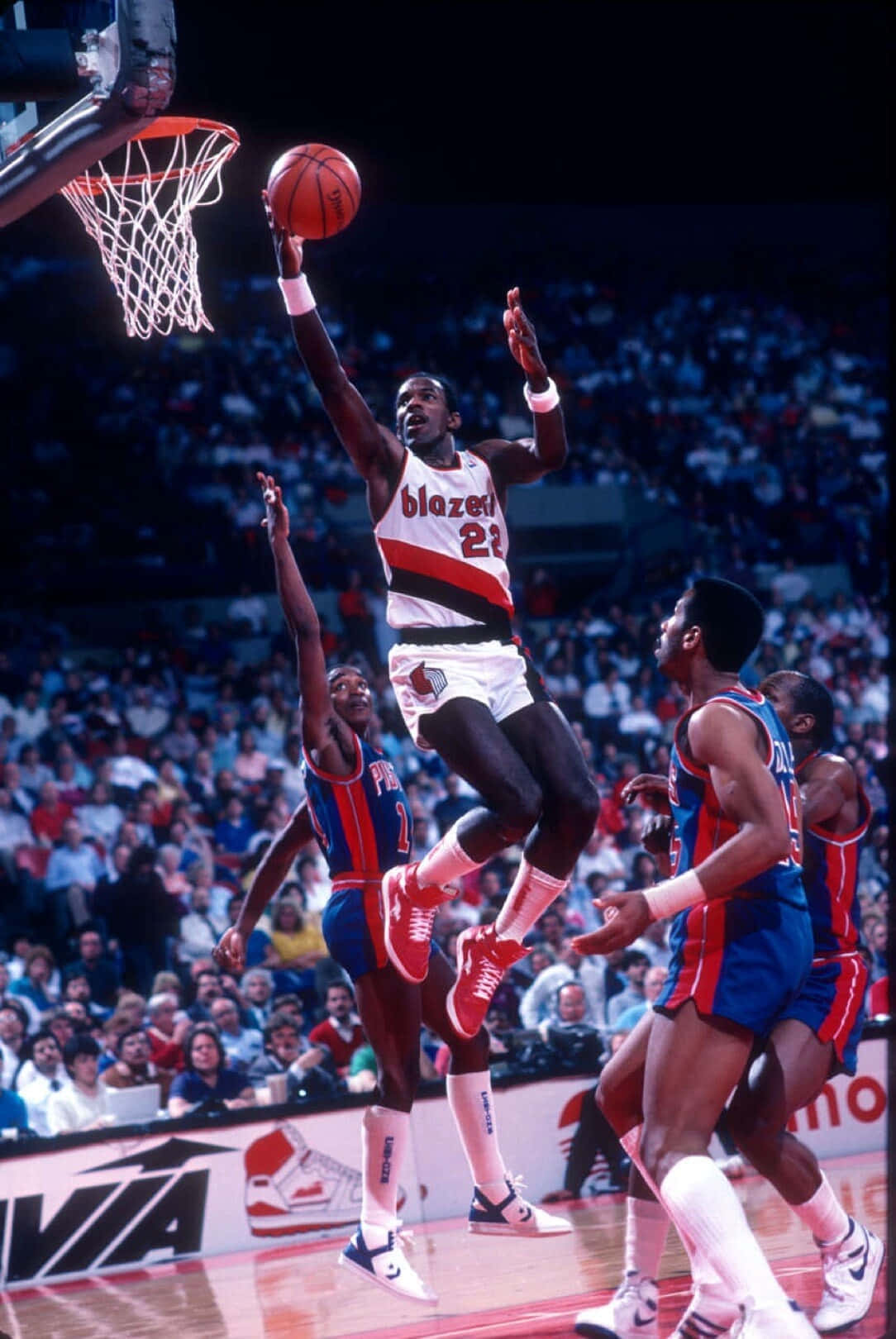 克萊德·德雷克斯勒1990年NBA總決賽開拓者隊 壁紙 — 一名身穿白色和紅色球衣的籃球運動員高高躍起扣籃，而身穿藍色球衣的防守球員在明亮的場館燈光下試圖封阻他。