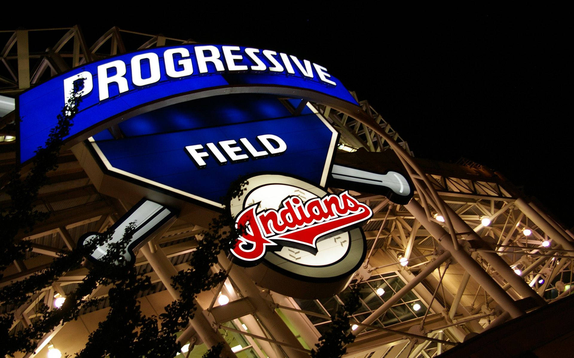 克里夫蘭印第安人隊普羅格雷西夫球場入口 壁紙 — 一個明亮照明的 Progressive Field 標誌，帶有 Indians 標誌，在黑暗的夜空下發光，由球場建築和樹枝框住。
