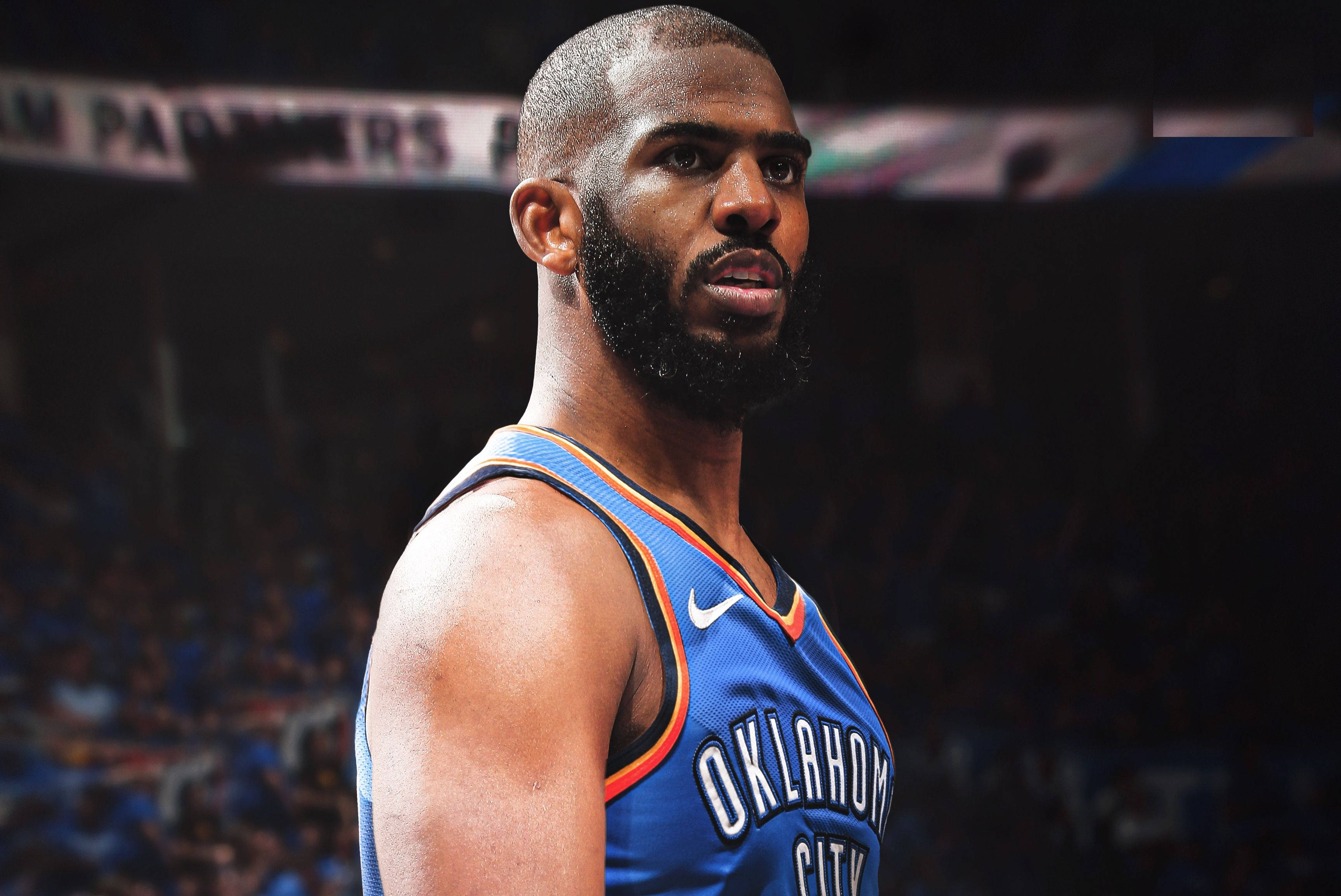 克里斯·保羅細節版OKC球衣 壁紙 — 一位專注的籃球運動員身穿奧克拉荷馬城球衣，站在燈光昏暗的球場上，身後是模糊的觀眾。