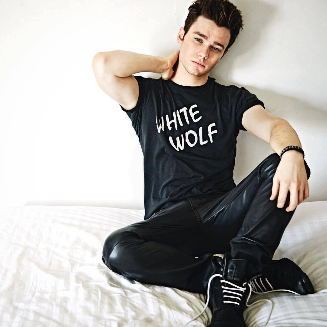 克里斯·柯爾弗時尚男裝拍攝 壁紙 — 一個穿著黑色「WHITE WOLF」T恤和皮褲的年輕男子，一手放在頸後，隨意地坐在白色床上。