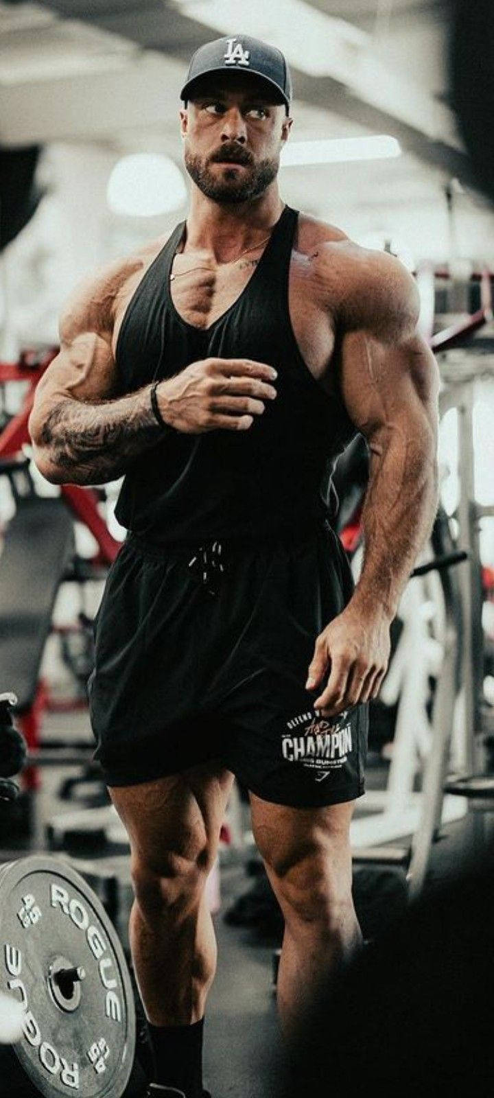 一名肌肉發達的男子在健身室戴著帽子和背心，舉起槓鈴擺姿勢。