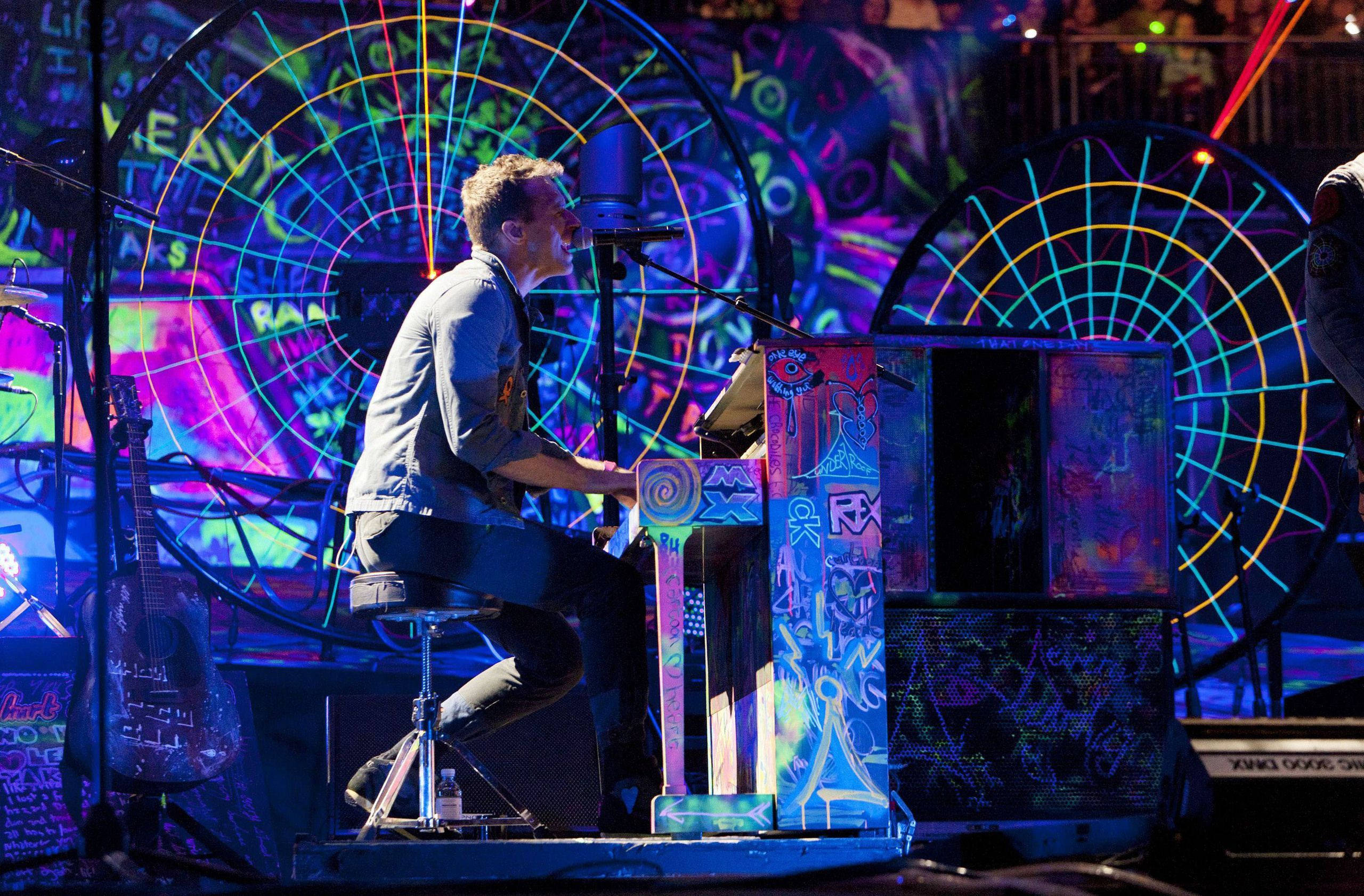 克里斯·馬汀 Mylo Xyloto 壁紙 — 一位音樂家在充滿活力的舞台上演奏塗鴉鋼琴，舞台以霓虹燈和圓形投影照亮。
