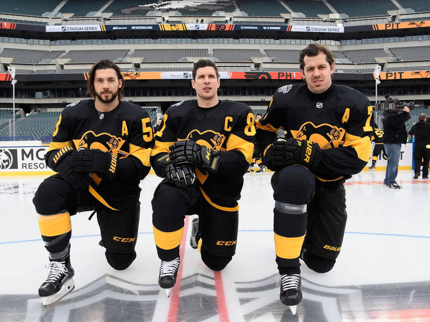 克里絲·萊唐2019 NHL Stadium Series 壁紙 — 三名身穿黑色和黃色球衣的曲棍球運動員跪在空蕩蕩的競技場冰面上，身後是體育場座位。
