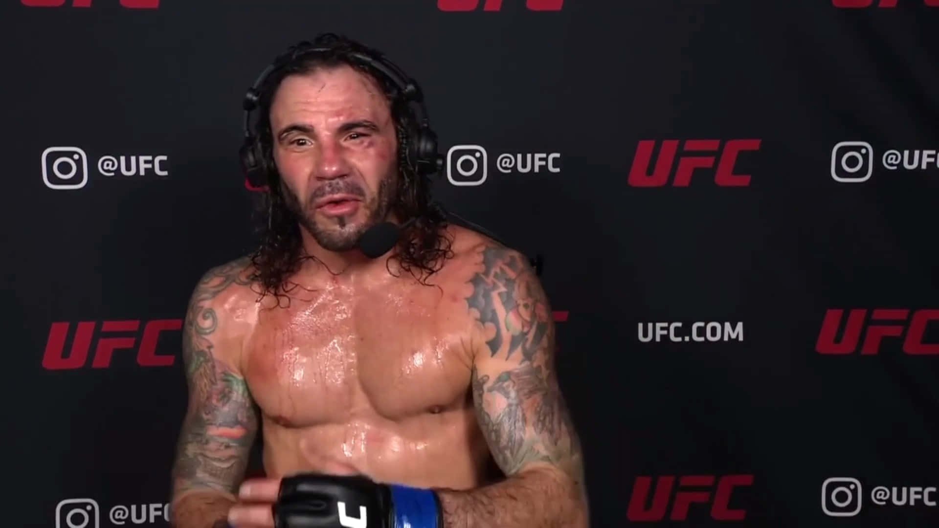 克雷頓·吉達對戰邁克爾·約翰遜於UFC Vegas 2021 壁紙 — 一個汗流浹背、有紋身的UFC拳手戴着耳機，在品牌黑色背景前對着咪高峰說話。