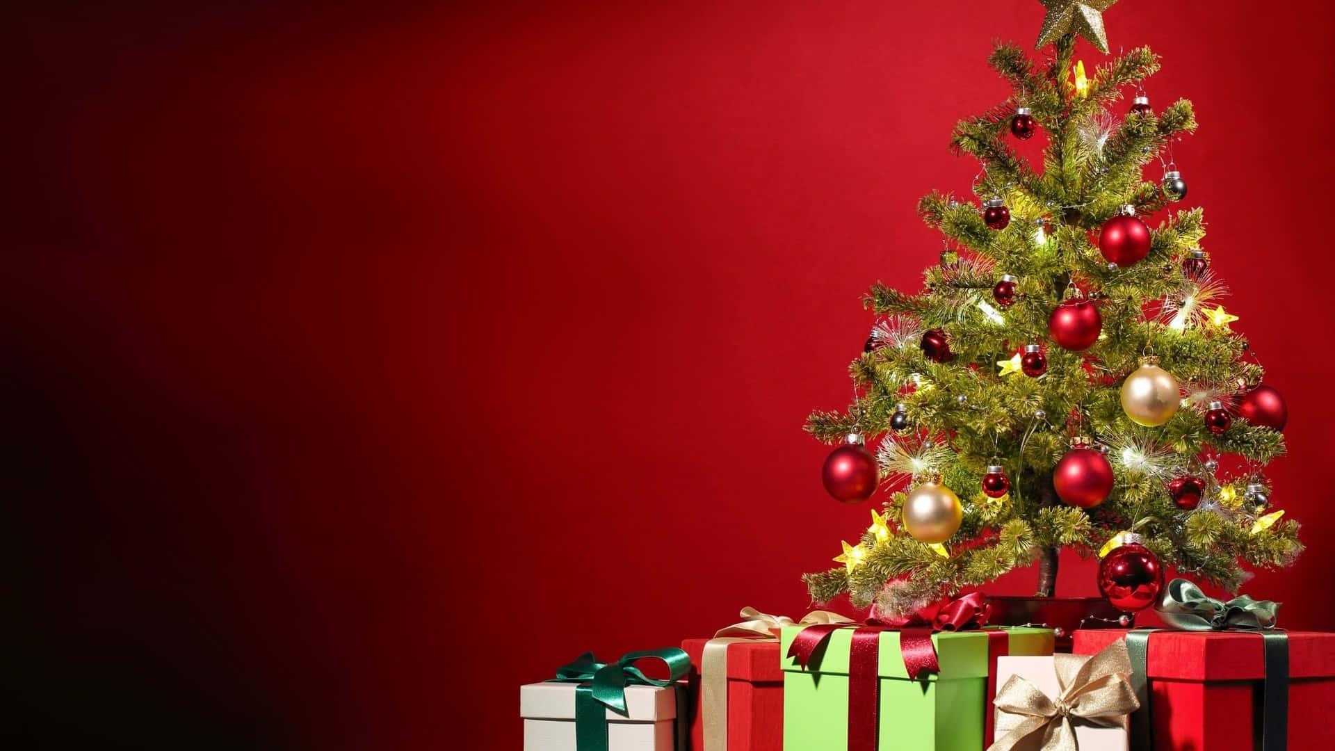 免費聖誕樹禮物圖片 壁紙 — 一棵節日聖誕樹裝飾著紅色、金色和銀色的飾物，旁邊放著色彩繽紛的禮物盒，背景是濃郁的紅色。
