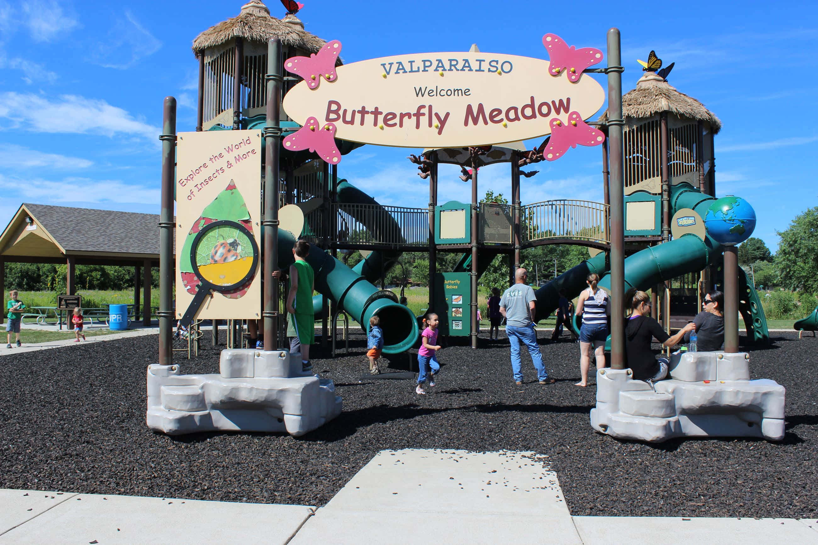 Butterfly Park 桌布 壁紙 — 兒童和成人享受一個充滿活力的綠色遊樂場，設有滑梯和蝴蝶主題標誌，在晴朗的藍天下。