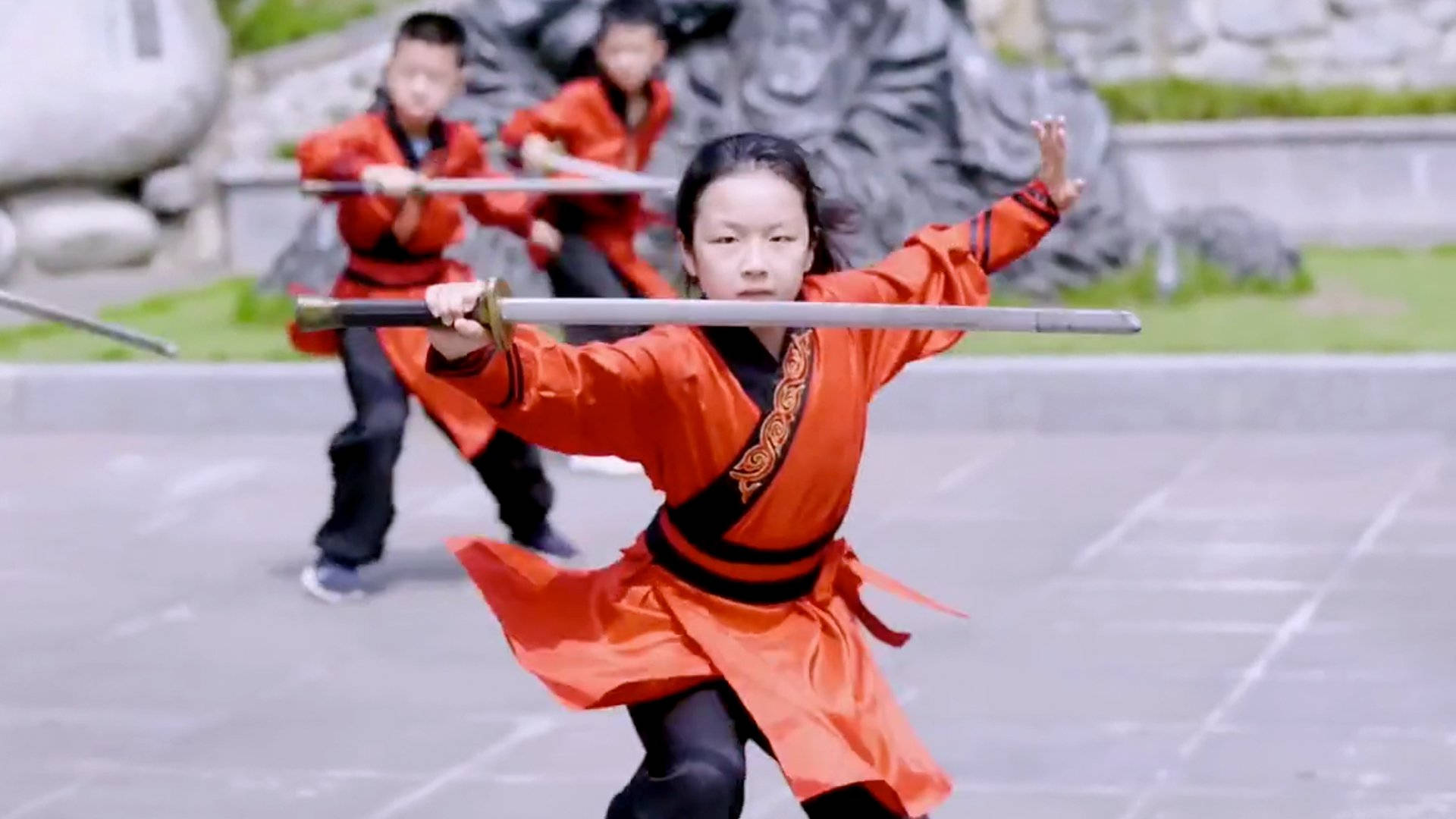 兒童武術功夫課 壁紙 — 一個年輕女孩穿著鮮豔的橙色武術制服，專注地揮舞著劍，而兩個男孩在她身後的一個石頭庭院裡訓練。