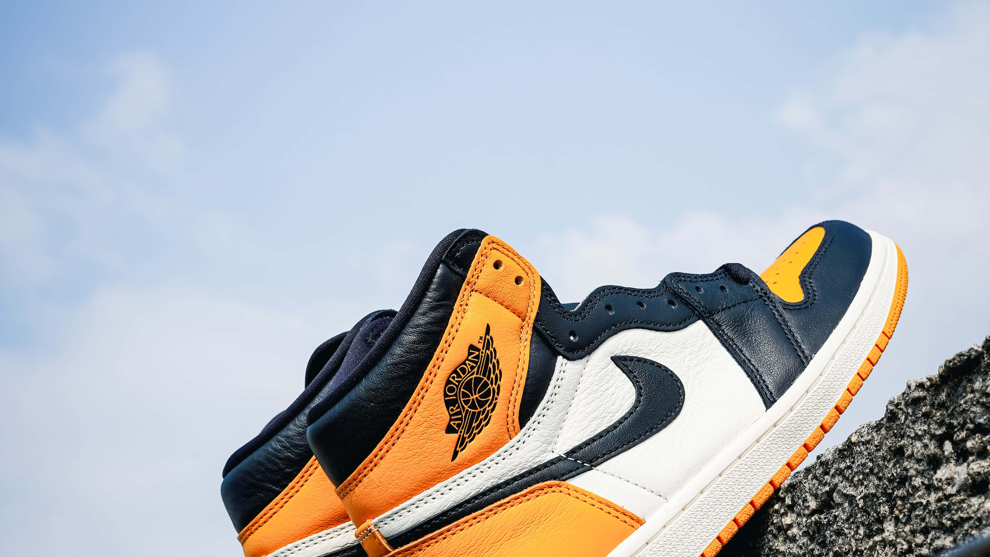 入手Air Jordan鞋款4K，打造舒適時尚造型 壁紙 — 海軍藍、橙色和白色的Jordan 1波鞋特寫，棲息在岩石表面上，襯托著明亮的藍天。
