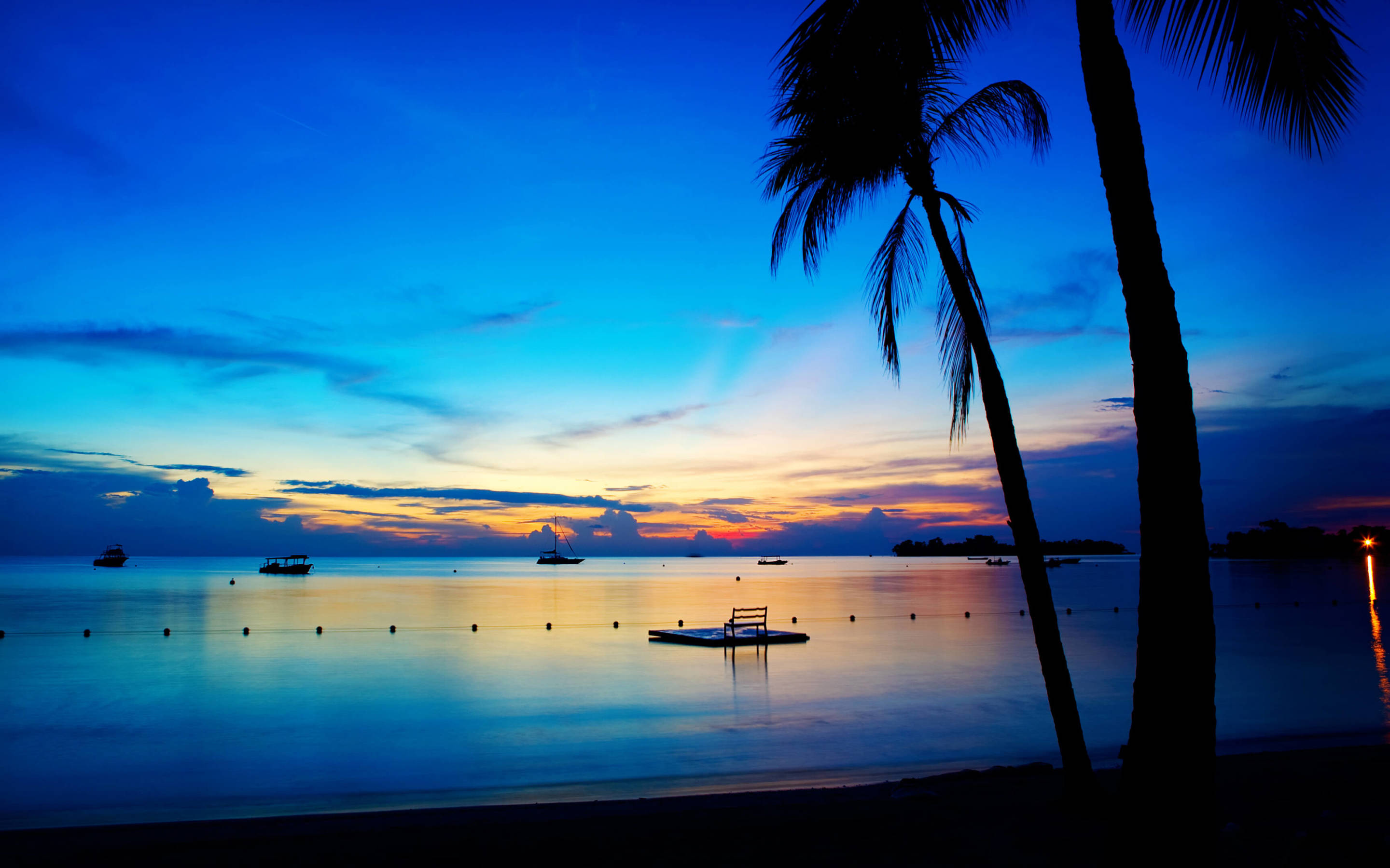 內格利爾七哩海灘夜景 壁紙 — 剪影的棕櫚樹框出一個寧靜的熱帶海灘，在黃昏時分，船隻漂浮在平靜的水面上，天空呈現深藍和暖橙色的漸變。