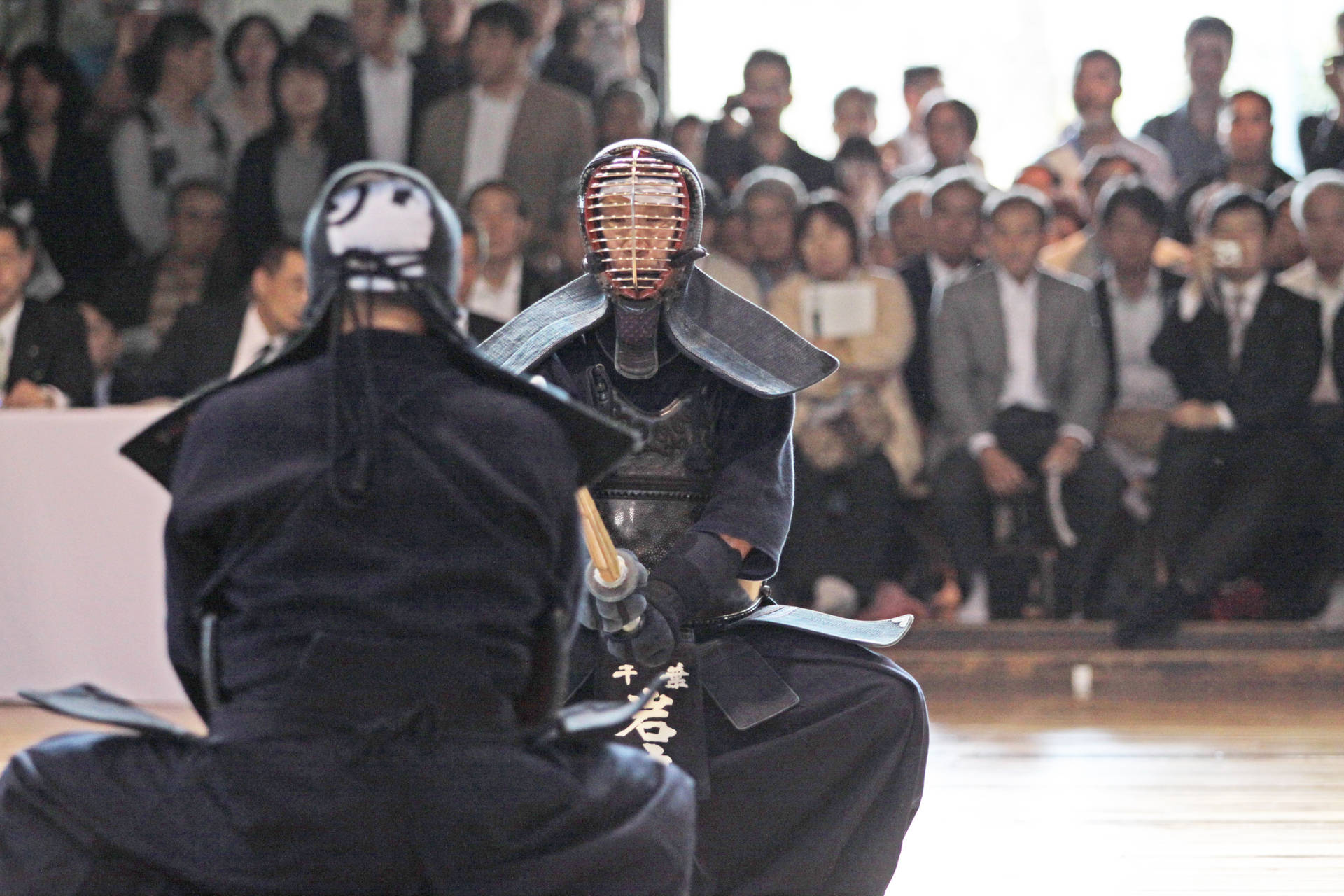 全副武裝劍道武士正面對峙 壁紙 — 兩名身穿全副盔甲的劍道戰士在木地板上對峙，背景中觀眾專注地觀看。