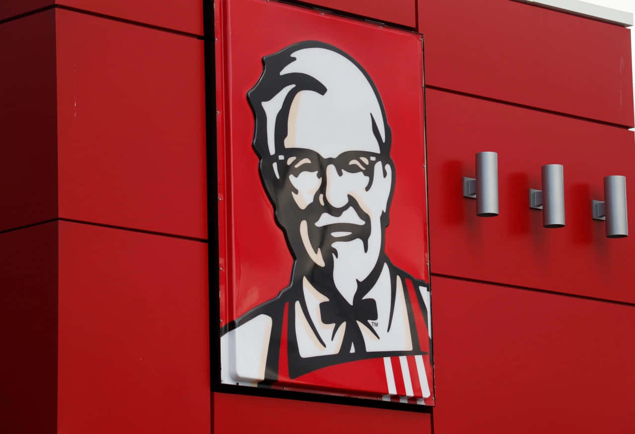 全家共享美味 KFC 餐點 壁紙 — 一座大膽的紅色KFC建築外牆，在明亮的日光下展示標誌性的山德士上校肖像標誌。