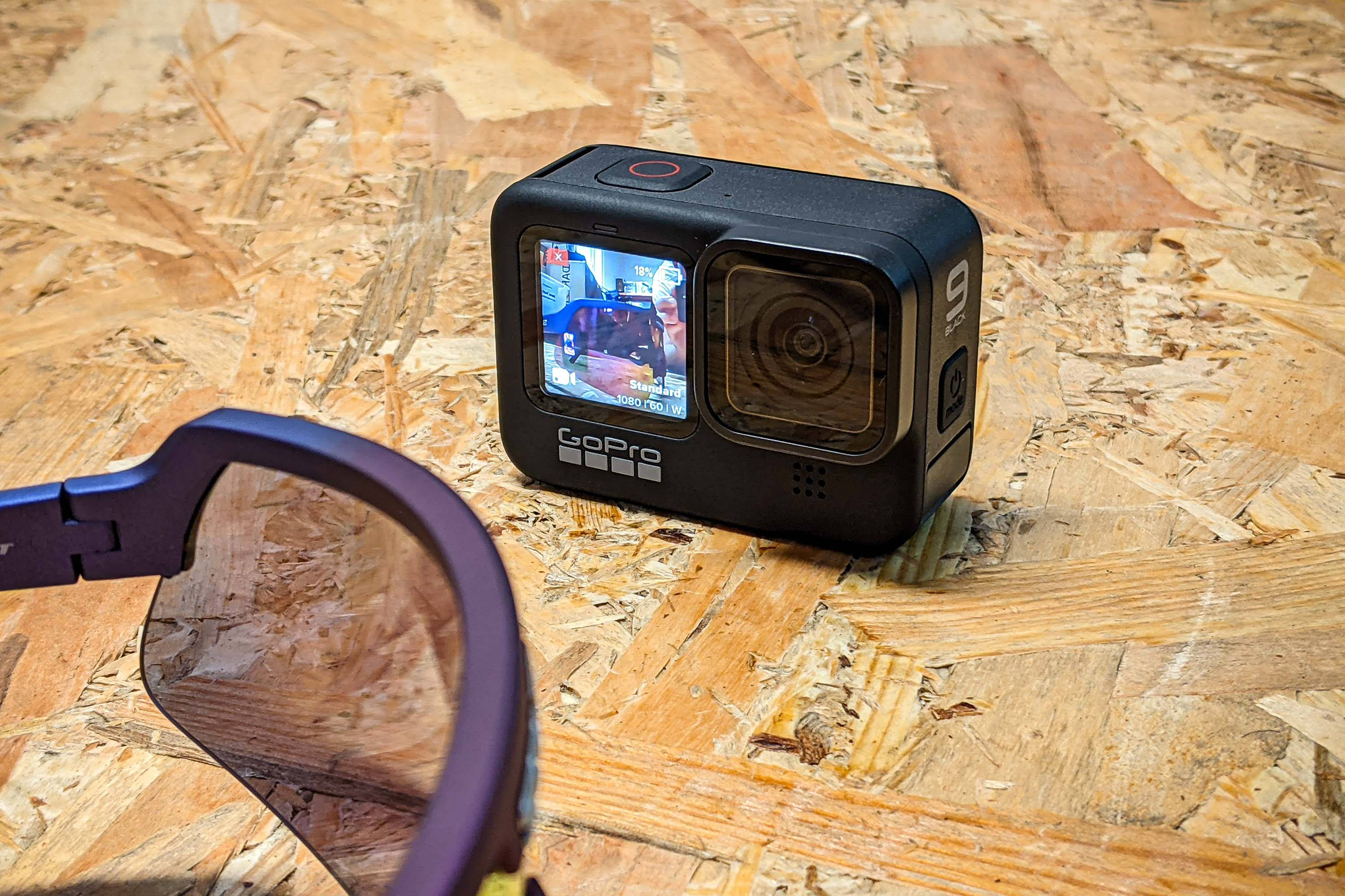 全新 GoPro Hero 9 精緻近攝圖 壁紙 — 一部黑色GoPro 9相機放在有紋理的OSB木板上，旁邊是紫色運動眼鏡，在明亮的日光下。