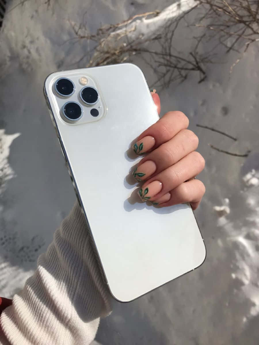 全新 iPhone 12 — 智能手機的未來 壁紙 — 一隻帶有葉子指甲藝術的手，在雪景背景中拿著一部白色智能手機，背景有光禿的樹枝。