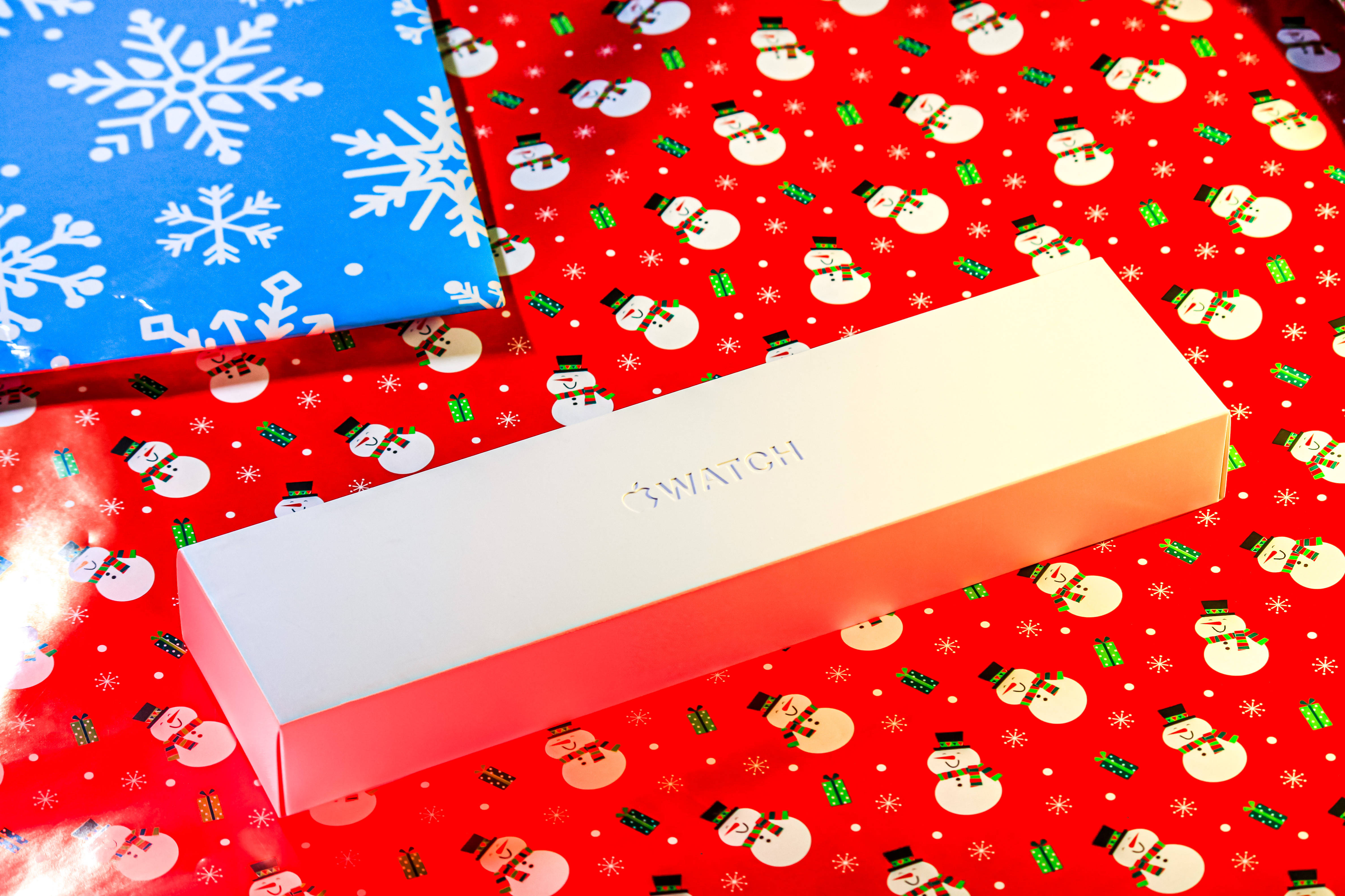 全新未拆封Apple智能手錶 壁紙 — 一個時尚的白色Swatch手錶盒放在節日紅色雪人包裝紙上，旁邊是藍色雪花包裝紙。