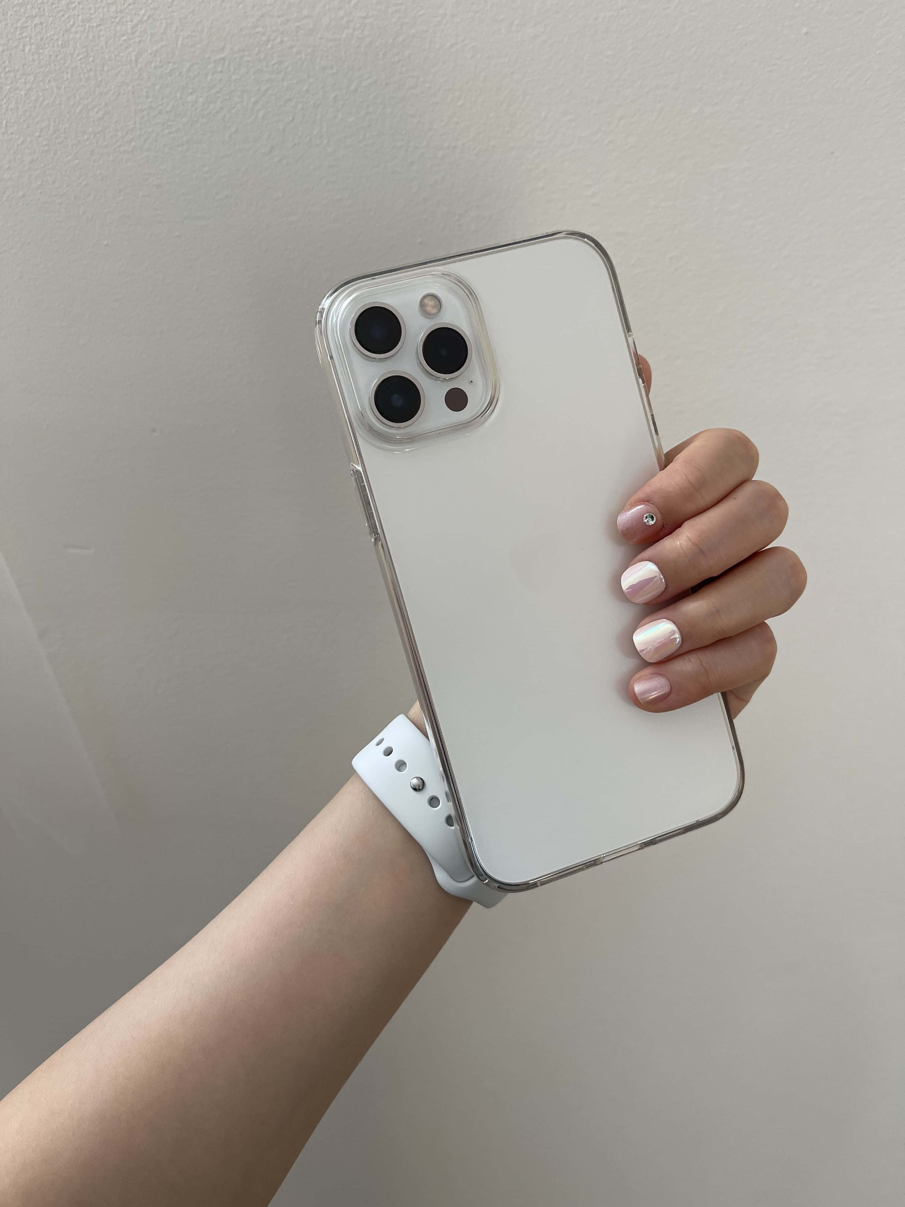 全新革命性 iPhone 12 壁紙 — 一隻帶有粉紅色閃粉指甲的手，拿著一個透明保護殼內的白色iPhone，背景是素色牆壁。