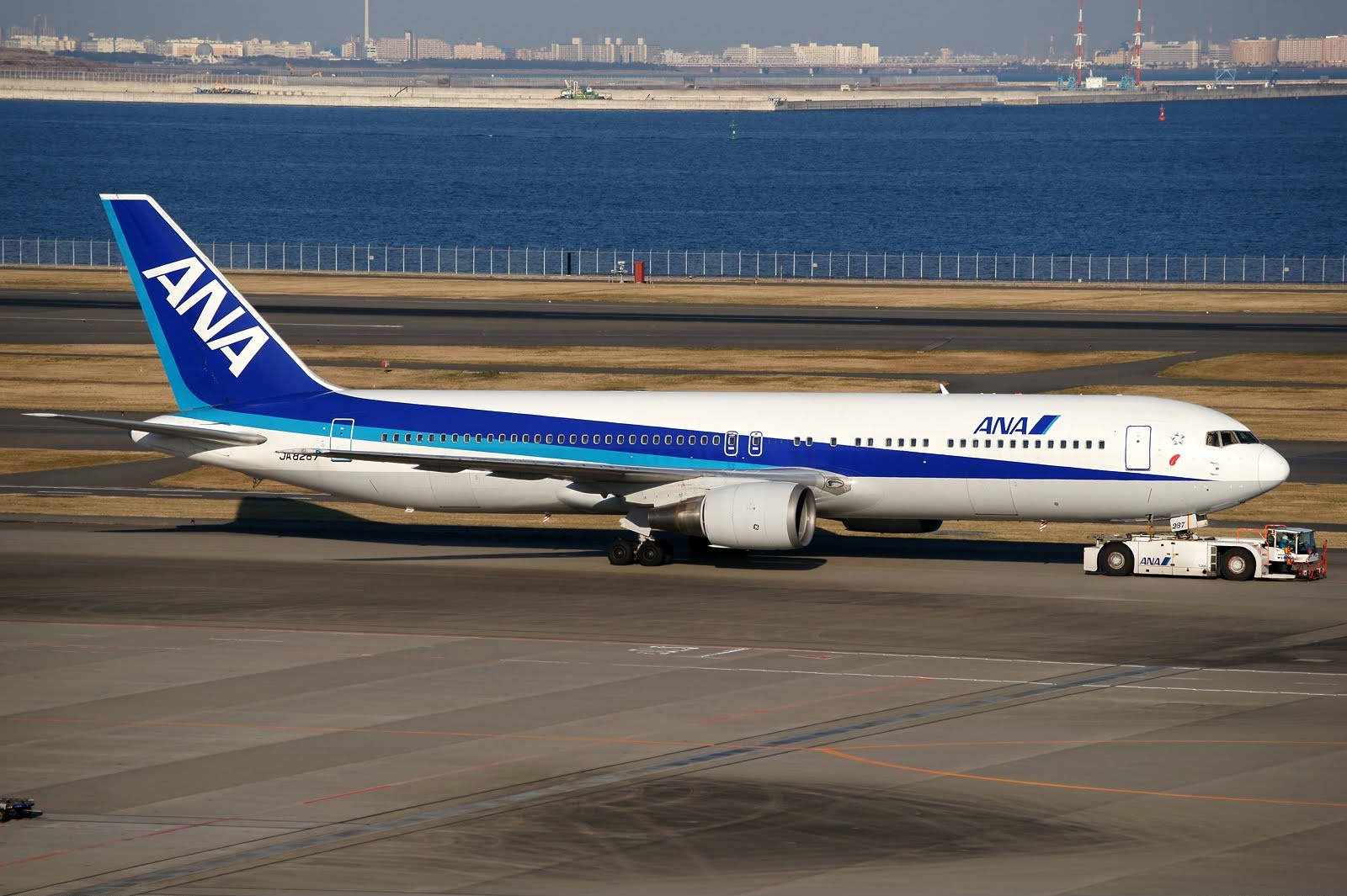 ANA飛機在機場停機坪上被拖行，靠近水和城市。