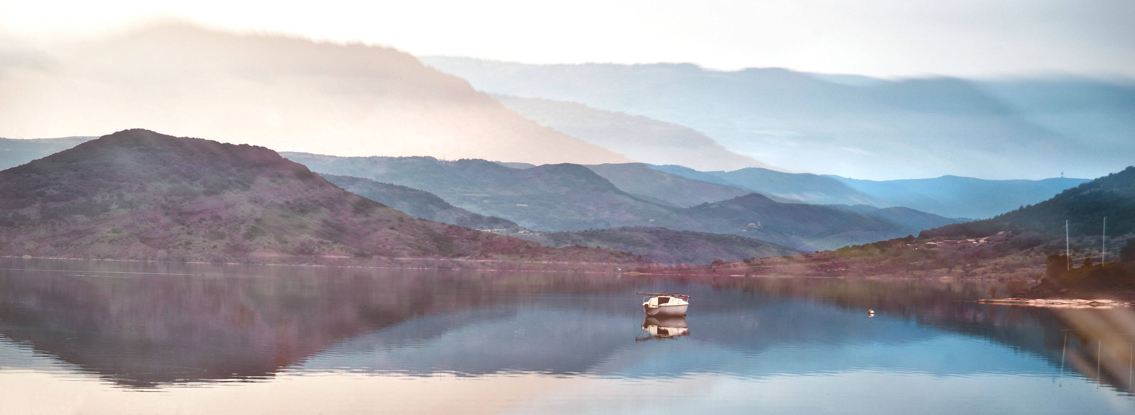 全景寧靜海景 壁紙 — 一艘孤獨的小船靜靜地漂浮在平靜的湖面上，映照著薄霧籠罩的山巒，在柔和的黎明天空下。