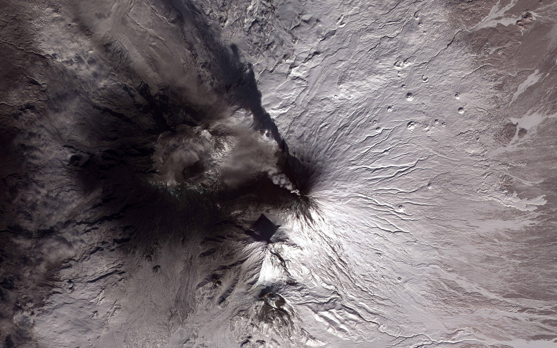 全景颶風 壁紙 — 白雪覆蓋的火山噴發，黑色火山灰羽狀物從火山口噴湧而出的航拍視圖。