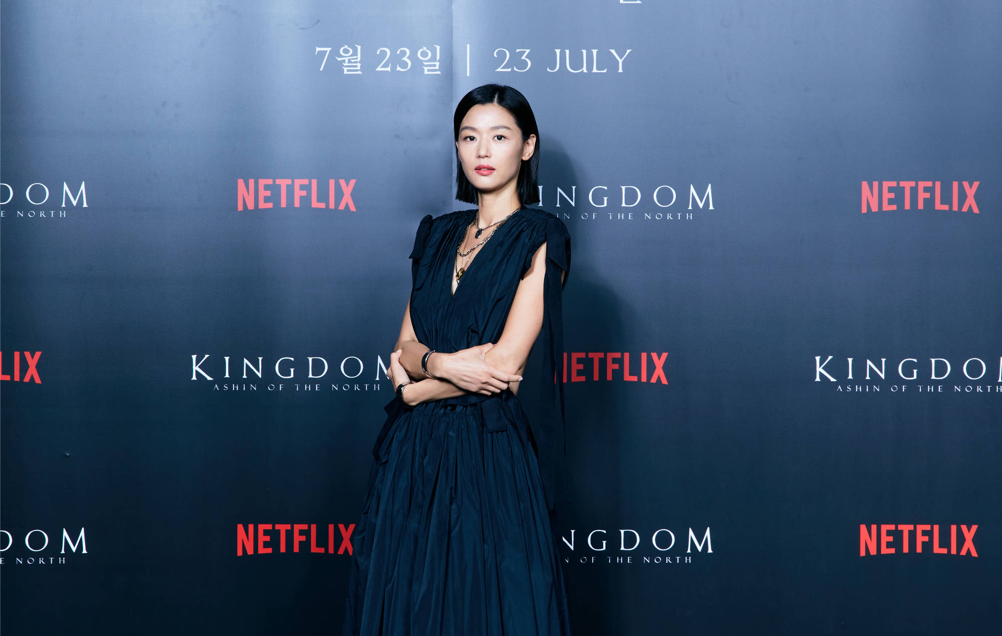 全智賢《王國》系列活動 壁紙 — 一位身穿深藍色禮服的優雅女士自信地站在Netflix Kingdom活動現場，身後有宣傳文字。
