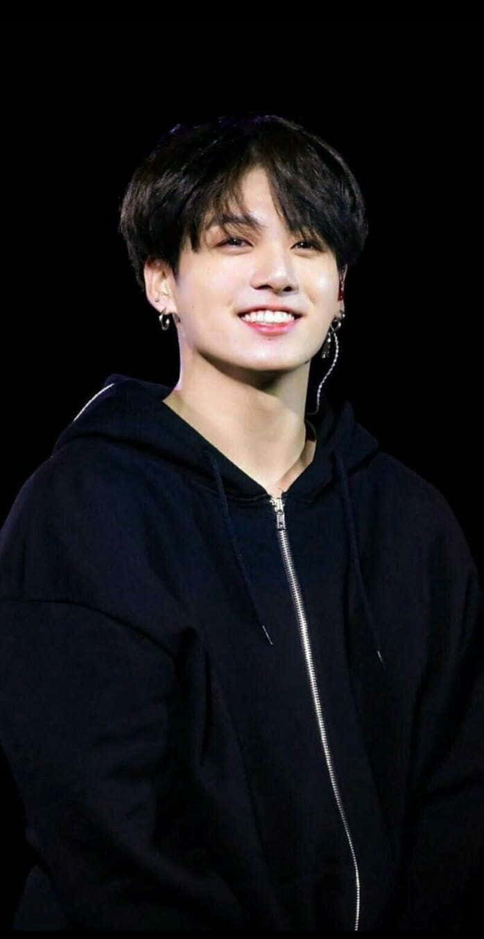全球巨星 Jungkook 的迷人肖像 壁紙 — 一位深色頭髮、戴耳環的年輕男子，在黑色背景前穿著黑色拉鍊連帽衫，溫暖地微笑。