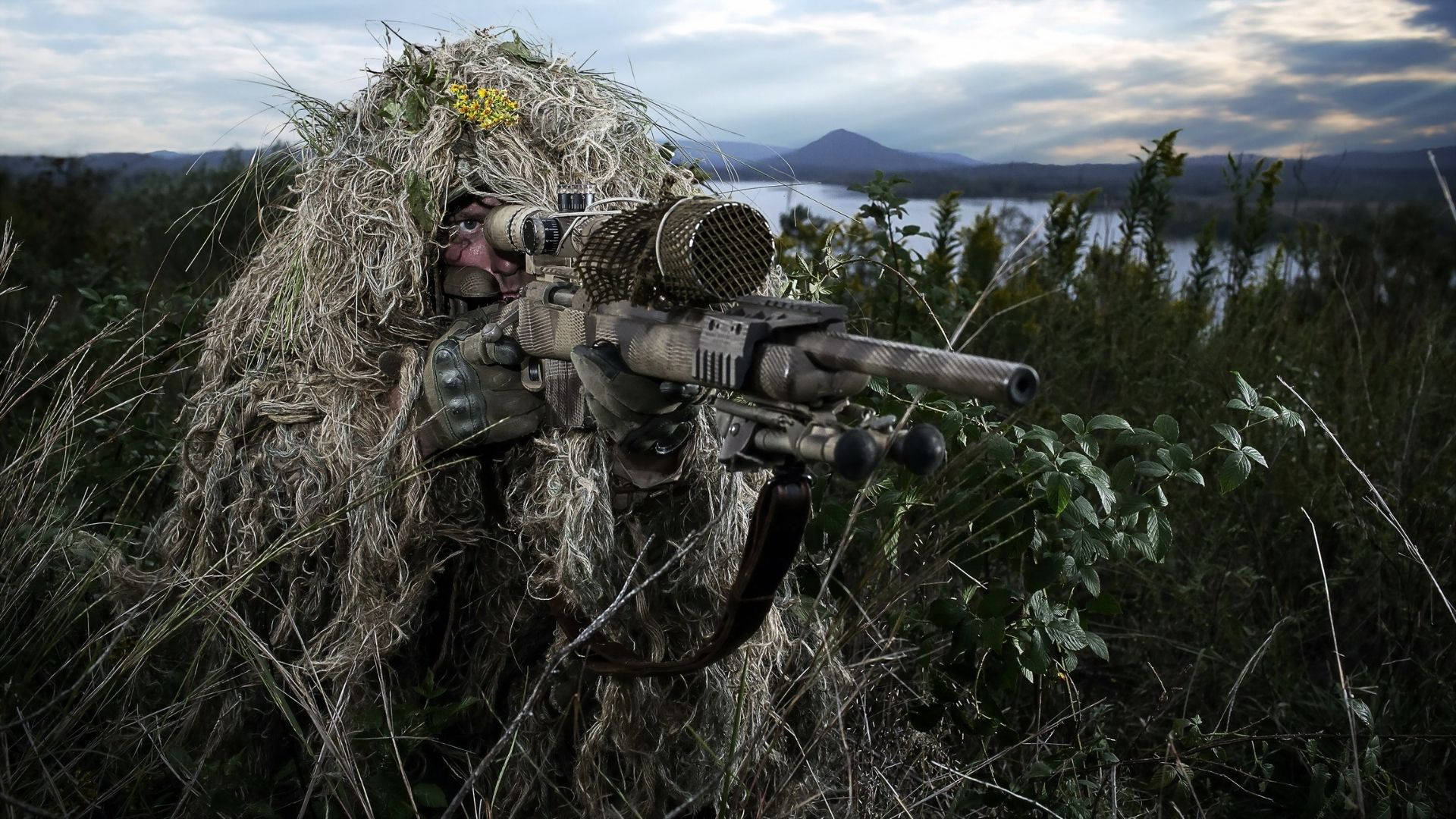 全裝備迷彩士兵持狙擊步槍 壁紙 — 一名穿著吉利服的狙擊手融入高草叢中，俯瞰著陰天天空下遠處山巒環繞的湖泊。