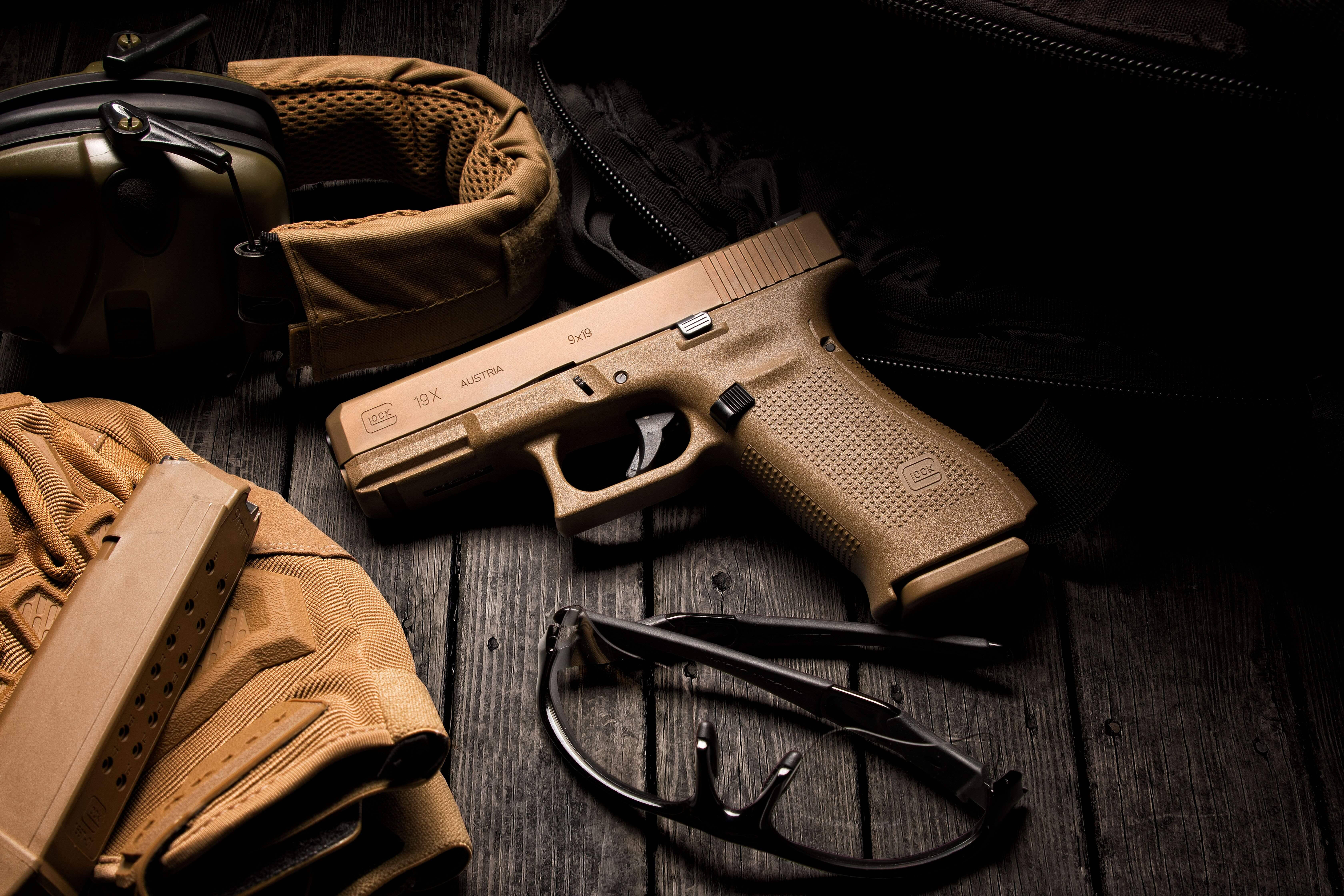 全銅色 Glock 19X 壁紙 — 一支棕褐色格洛克手槍放在風化木上，旁邊有戰術裝備和護目鏡，在戲劇性的低調燈光下。