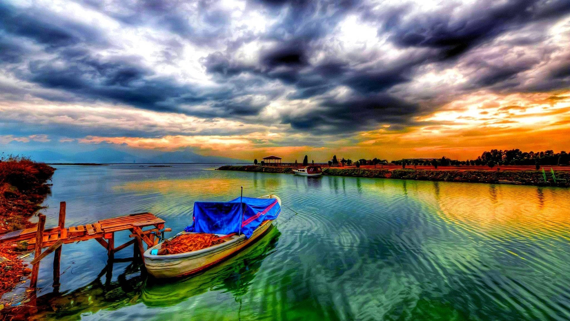 一艘帶有藍色防水布的小船停在木碼頭旁，色彩繽紛的日落倒映在水面上。
