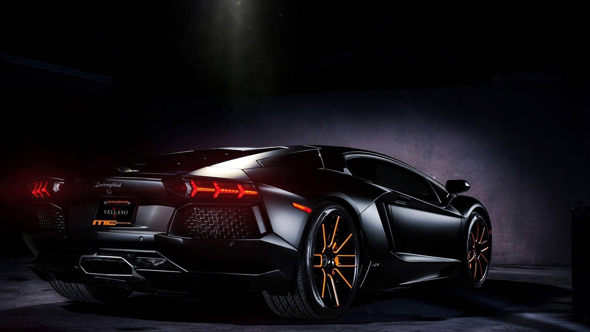 全高清黑色蘭博基尼跑車 壁紙 — 一輛啞光黑色的林寶堅尼Aventador，帶有發光的橙色輪圈和尾燈，在黑暗的攝影棚中戲劇性地照亮。