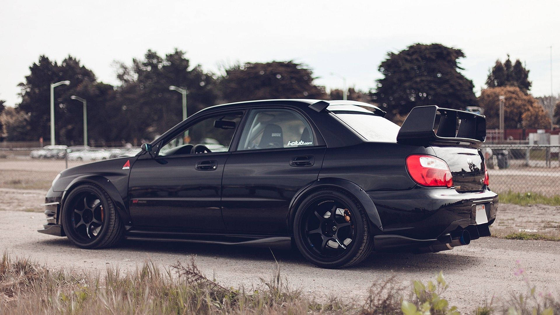 全黑塗裝斯巴魯車 壁紙 — 一架時尚的黑色 Subaru Impreza WRX STI 配備大型後擾流板，停泊在碎石地上，周圍樹木環繞，天空多雲。