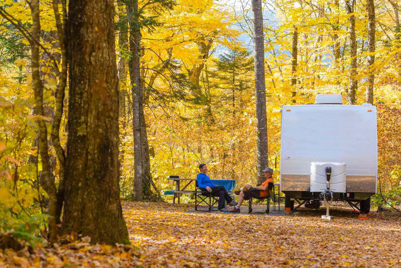 Fall Camping 桌布 壁紙 — 兩人在金色秋葉燦爛的森林中，在白色露營拖車旁的摺疊椅上放鬆。