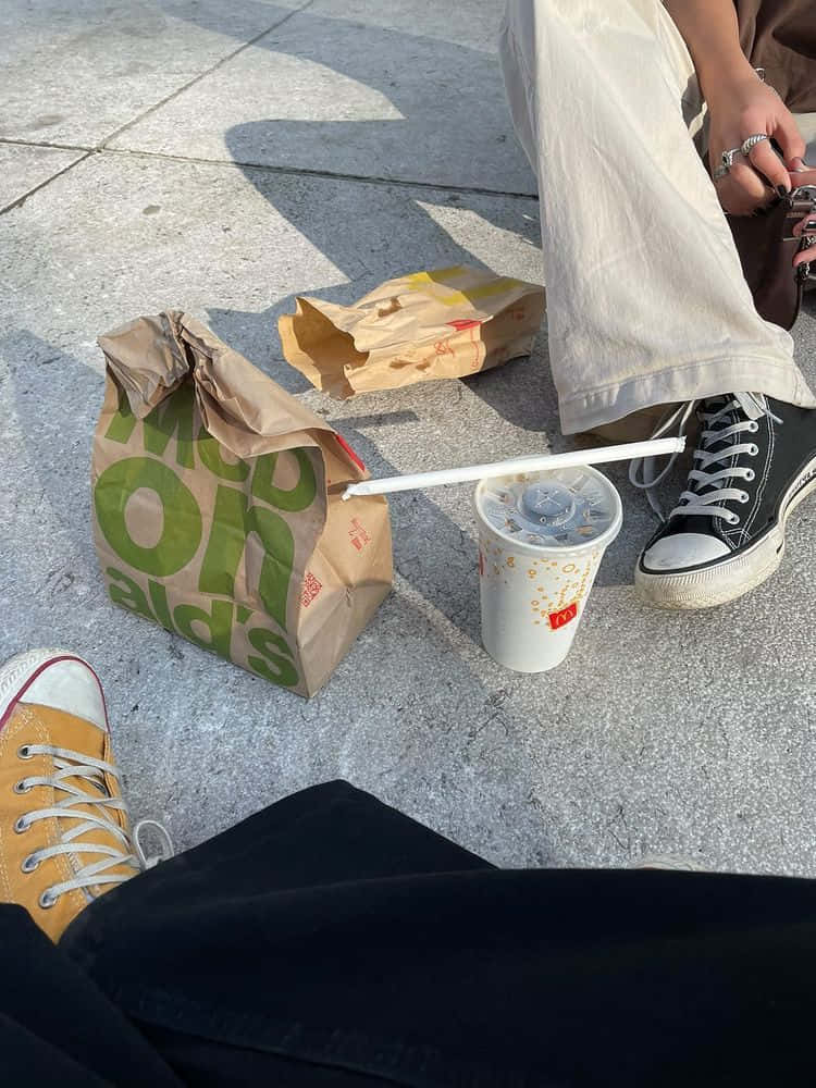 兩人坐地享麥當勞餐點圖片 壁紙 — 兩個人坐在行人路上享受麥當勞的食物和飲品，旁邊有運動鞋和紙袋。