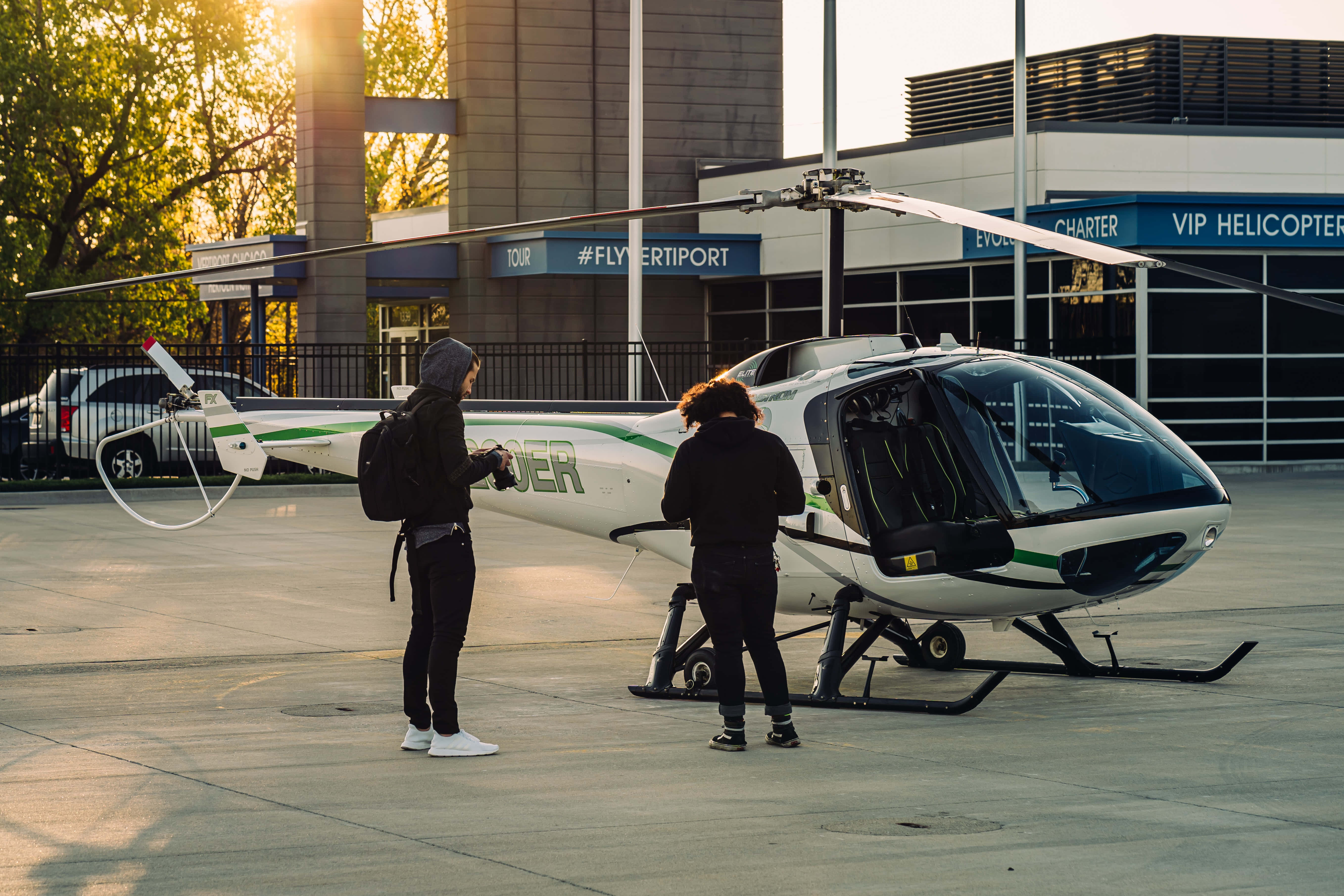 直升機 桌布 壁紙 — 兩人站在VIP直升機停機坪的一架白色直升機旁，金色陽光穿過樹林灑落。