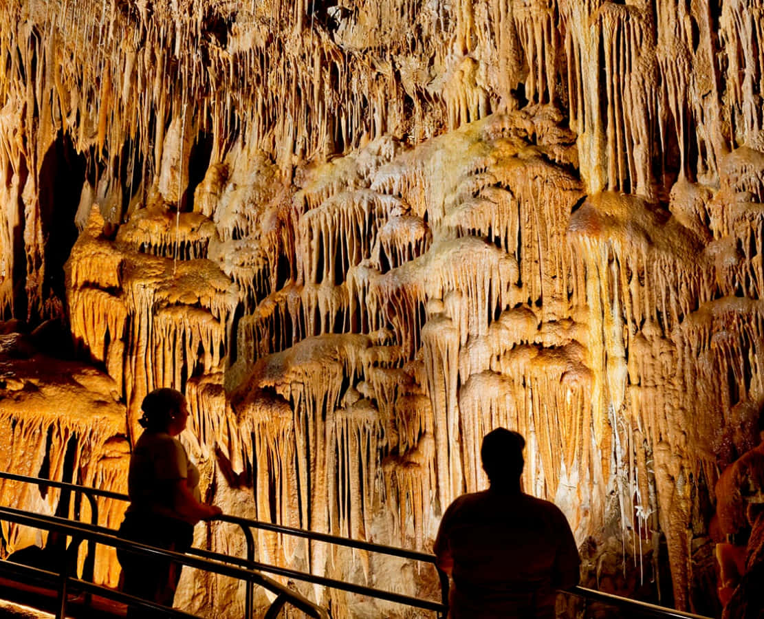Cave 桌布 壁紙 — 兩位剪影遊客在溫暖燈光的洞穴中，敬畏地凝視著高聳的金色鐘乳石和石筍。