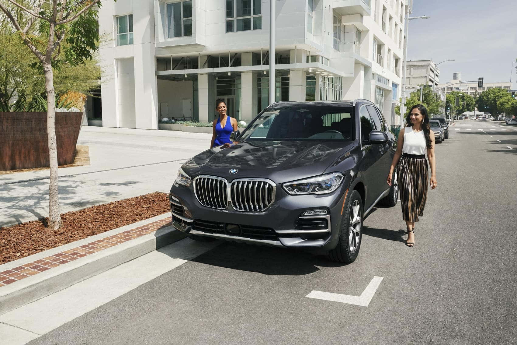 Bmw X5 桌布 壁紙 — 兩位女士喺陽光明媚嘅城市街道上，喺一架停泊嘅時尚灰色BMW SUV旁邊散步，周圍有現代建築同樹木。